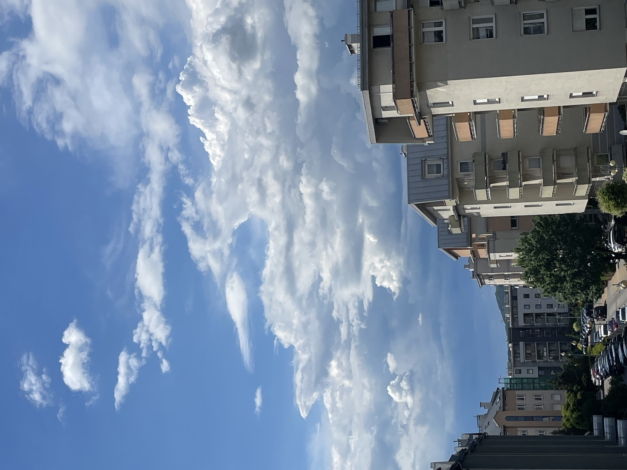 This image captures a daytime urban landscape in the city of Unknown, Unknown, from an elevated vantage point. The scene is dominated by residential apartment buildings and a partially cloudy sky.
The upper two-thirds of the image display a bright blue sky transitioning into a mixture of white and grey cumulus clouds. The top-left features clear blue sky with smaller, scattered white clouds, while the central and right portions exhibit larger, denser formations of white and dark grey clouds, indicating varying cloud cover.
In the foreground and mid-ground, numerous multi-story apartment buildings are visible. The buildings on the right side are primarily light beige or grey with distinct dark grey sloped roofs and multiple rectangular balconies featuring light brown or orange railings. These structures have numerous rectangular windows. A similar style of building is partially visible on the far left. Further into the scene, additional apartment complexes with varying architectural styles and colors line a curving street.
The street below is densely packed with parked vehicles, predominantly dark-colored cars, arranged along both sides. Additional cars are visible in motion on the street. Trees with green foliage are interspersed among the buildings, particularly a large deciduous tree on the right side of the street. Streetlights are also discernible along the road. In the far distance, hills or mountains are faintly visible beneath the cloud line. No people are explicitly identifiable in the image.