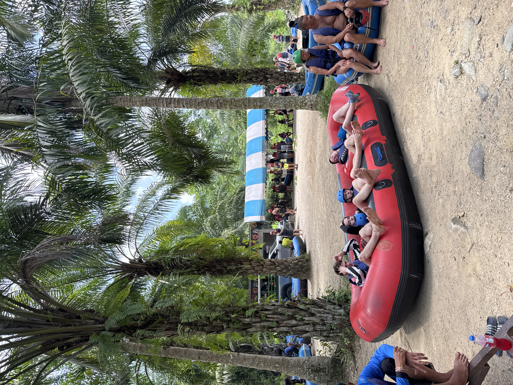 The image shows a scene in Tambon Song Phraek, Thailand. Several rafts are present on a dirt or sandy surface, with people relaxing in them. Some individuals are wearing helmets and life jackets, suggesting a water activity like rafting. Other people are standing and sitting around the rafts. Palm trees are prominent in the foreground and background. A blue and white striped structure is also visible in the background. The environment appears to be a recreational area near a river or body of water.
