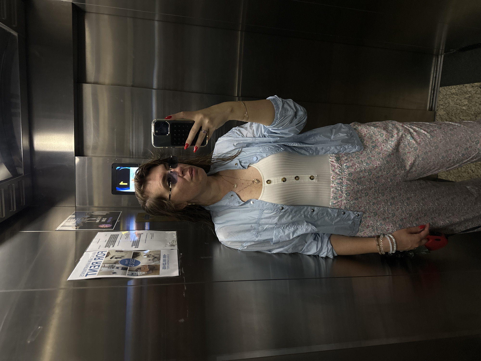 A young woman with long brown hair and sunglasses is taking a mirror selfie in an elevator. She is wearing a white ribbed tank top, a light blue button-up shirt, and floral patterned pants. She has red nail polish and is wearing bracelets on her left wrist. A red object, possibly a phone case, is visible in her hand.
The elevator has stainless steel walls and doors. There are a few posters on the elevator wall. One poster is for "FOR RENT" and displays a phone number and what appears to be a website or company name. The lighting suggests it is daytime. The woman's expression is neutral. The setting is an interior space, likely an apartment building or hotel, given the elevator and the "FOR RENT" sign. The location context provided is Batumi, Georgia.