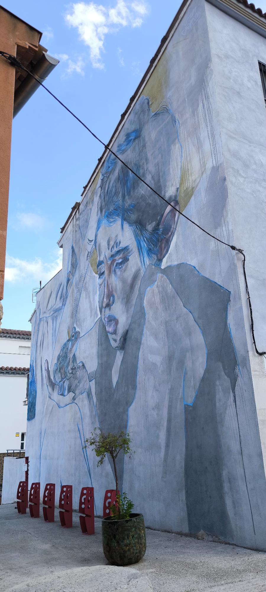 The image shows a mural on the side of a building in Fanzara, Spain. The mural depicts a large, stylized human face with blue accents in the hair and eyes, rendered in a muted color palette of grays and yellows. The subject is looking down, with its mouth slightly open. In its outstretched hand, the mural shows a frog with a small crown on its head. Below the mural, there are several red, stylized benches arranged in a row, and a small tree in a dark, speckled pot. The building facade is textured and appears to be made of concrete. The sky above is blue with some scattered white clouds. Electrical wires run across the mural. The overall scene is urban, with architectural elements from adjacent buildings visible.