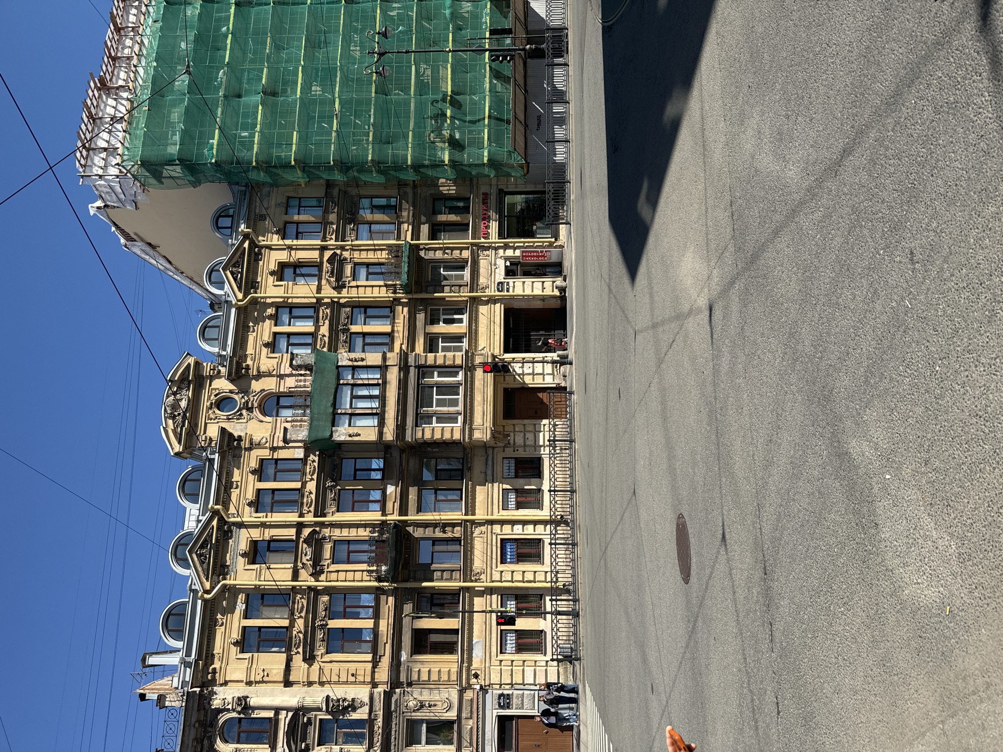This image captures a view of a street in Saint Petersburg, Russia, on a clear, sunny day. The focal point is a multi-story yellow building with ornate architectural details and arched windows. To the right of this building, a section of an adjacent structure is under renovation, covered in green scaffolding and netting. The street in the foreground is paved and appears to be a crossroads, indicated by a pedestrian crossing and traffic lights. Several people are visible on the sidewalk in front of the yellow building, and one person is walking on the street. Power lines crisscross the sky. Shadows cast by the buildings and utility poles stretch across the asphalt.