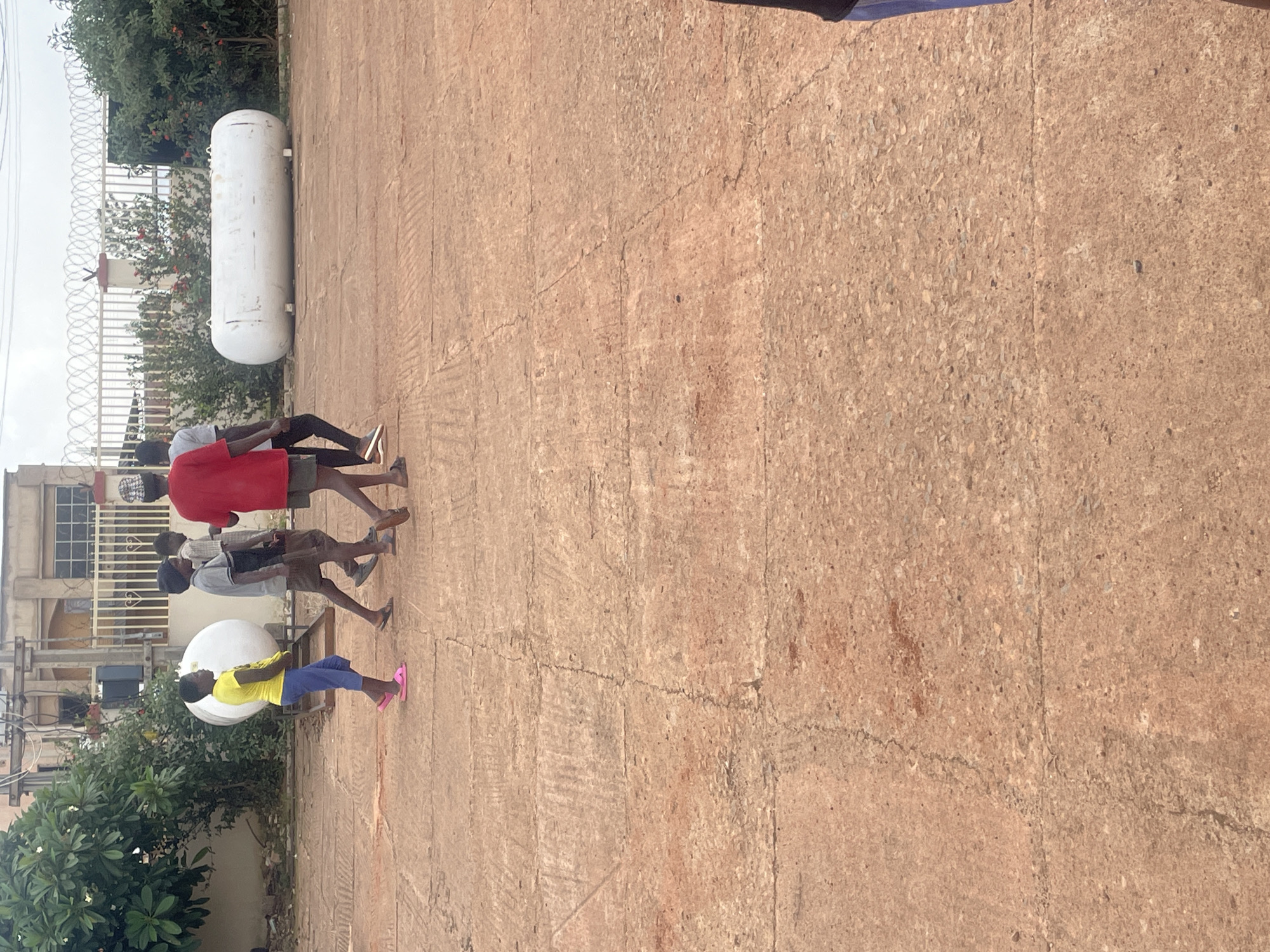 A group of four children are walking across a paved area in Jos, Nigeria. The children are of varying ages and are dressed casually. The ground is made of large, rectangular concrete slabs with visible cracks and wear. In the background, there is a white cylindrical tank, a metal fence with barbed wire, lush green trees, and a building. The sky is overcast. The image appears to be taken during the day.
