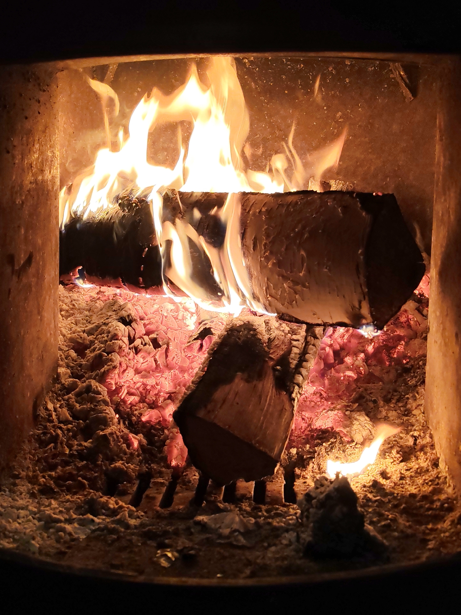 The image shows the interior of a fireplace or wood-burning stove. Flames are visible burning on and around two large logs in the upper portion of the image. Below the logs, there is a bed of glowing red and orange embers mixed with ash. At the bottom center, there is another small log partially buried in the ashes and embers, along with what appears to be a lump of ash or unburnt fuel. The interior walls of the fireplace are visible on either side, showing a rough, textured surface. The image does not provide sufficient information to confirm its location in Chaikovsky, Russia, or to identify any people or specific interactions.