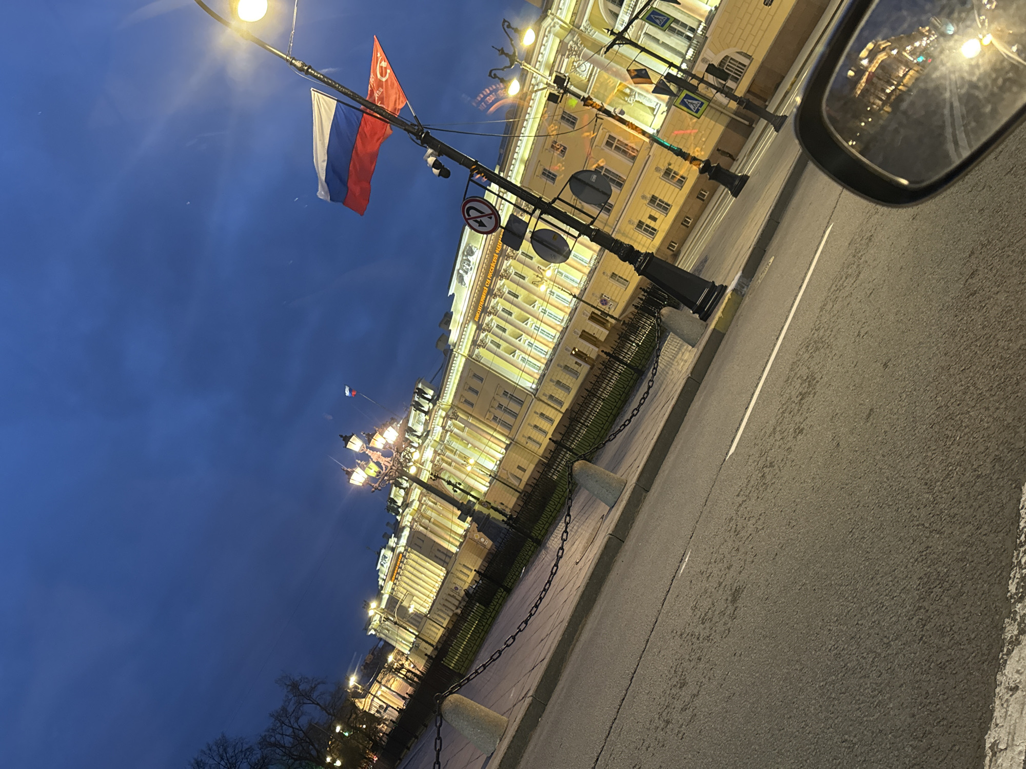 An urban scene in Saint Petersburg, Russia, is captured at dusk or twilight, viewed from the interior of a vehicle as indicated by the prominent reflection in a side mirror on the right. The sky is a deep, clear blue, contrasting with the warm, golden illumination of numerous streetlights and a grand, classical building.
The impressive multi-story building, likely the main building of the Russian State Pedagogical University named after A.I. Herzen (visible text: "РОССИЙСКИЙ ГОСУДАРСТВЕННЫЙ ПЕДАГОГИЧЕСКИЙ УНИВЕРСИТЕТ"), dominates the background. Its facade features columns and numerous windows, all brightly lit.
In the mid-ground, a paved road with a white lane marking stretches across the image. Alongside it, a sidewalk is delineated by a curb, and a series of concrete bollards connected by chains provide a barrier. Decorative black lampposts with multiple glowing fixtures stand along the sidewalk. Overhead, another lamppost extends across the street, from which two flags are flown: the white-blue-red national flag of Russia and a red flag bearing a yellow hammer and sickle emblem, characteristic of the Soviet era. Several traffic signs, including a "No U-turn" sign, are also attached to the lampposts. Bare tree branches are visible in the lower-left corner. The overall mood is quiet and atmospheric, highlighting the city's architectural beauty under artificial light.