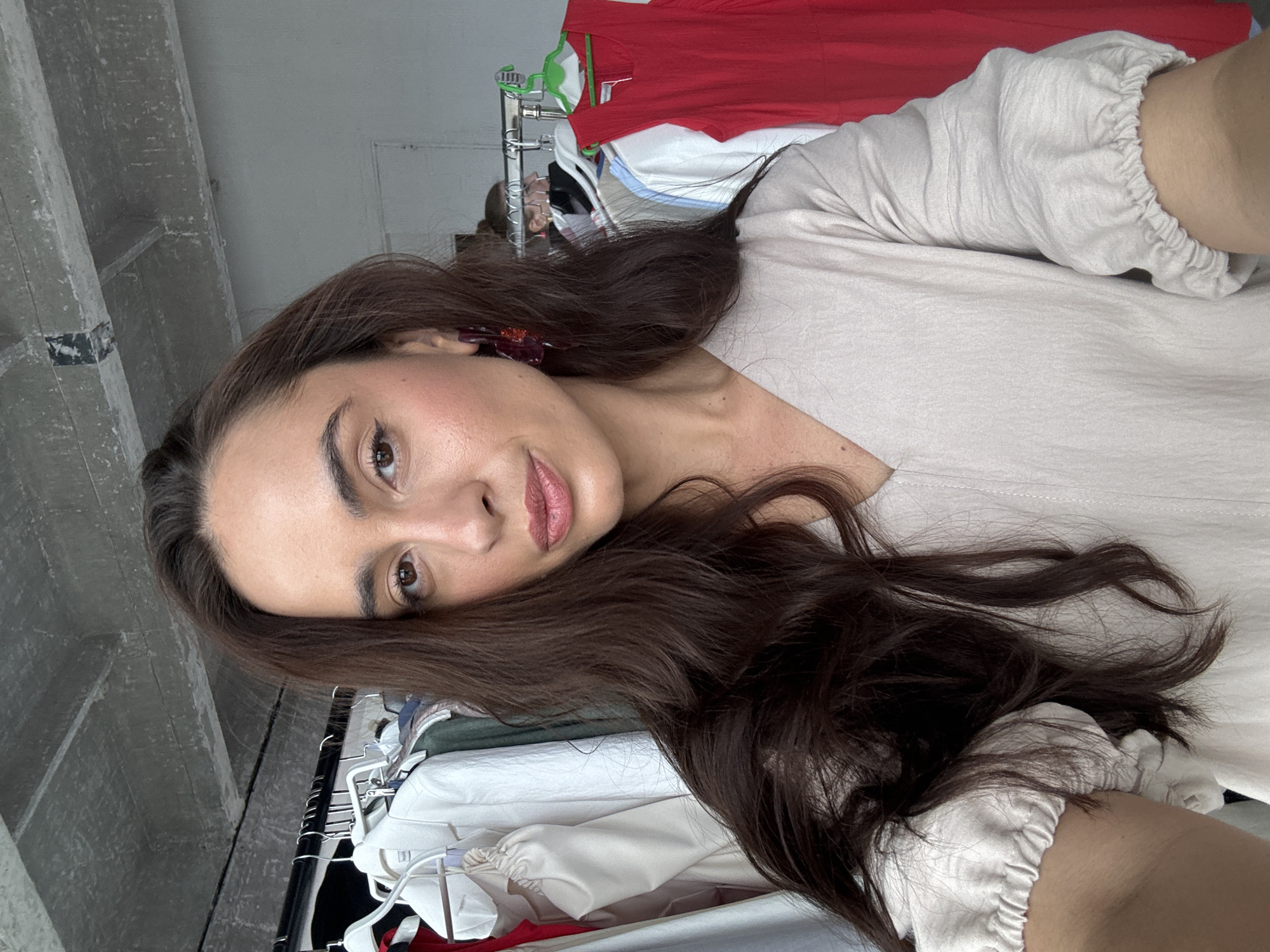 A medium shot captures a woman with long, dark brown hair and brown eyes looking directly at the camera. She is wearing a light-colored, V-neck top with puffed sleeves. Visible in the background are clothing racks filled with various garments, including white jackets, a red dress, and other clothing items, suggesting a retail or studio environment. The setting appears to be indoors, with a concrete ceiling and white walls. No specific location in Krasnoyarsk, Russia, is indicated.