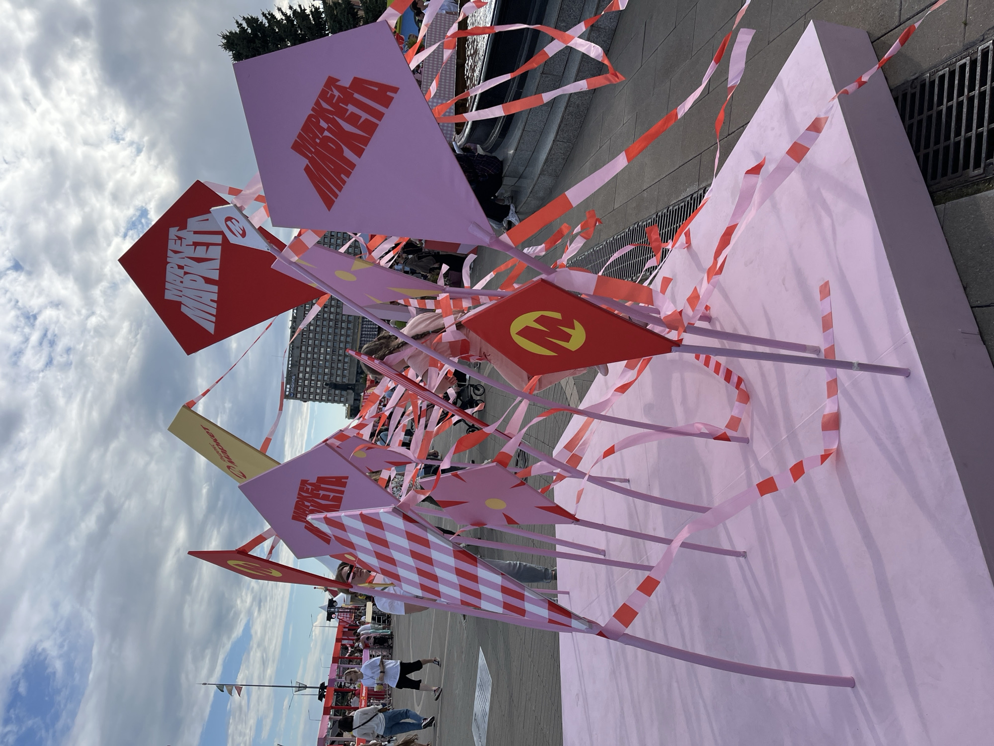 This is a medium shot, taken outdoors on a cloudy day, of a public art installation in Nizhny Novgorod, Russia, near the Volga River embankment. The installation consists of several large, diamond-shaped flags of varying colors (pink, red, yellow) with the stylized Russian word "МАРКЕТ" (MARKET) printed on them in bold, red, sans-serif font. Red and pink striped tape is draped and tied around the flags and supporting poles, creating a festive, almost celebratory atmosphere. Some of the flags also feature a circular logo with the letter "M" inside a yellow diamond.
In the background, several people are visible, casually dressed and walking around. Some are looking at the installation, while others are engaged in conversation or walking away. A tall, modern building and a church steeple are visible in the far distance, as is a large evergreen tree. The ground is paved with gray tiles, and a metal grate is visible in the foreground. The overall impression is that of an outdoor event or festival.