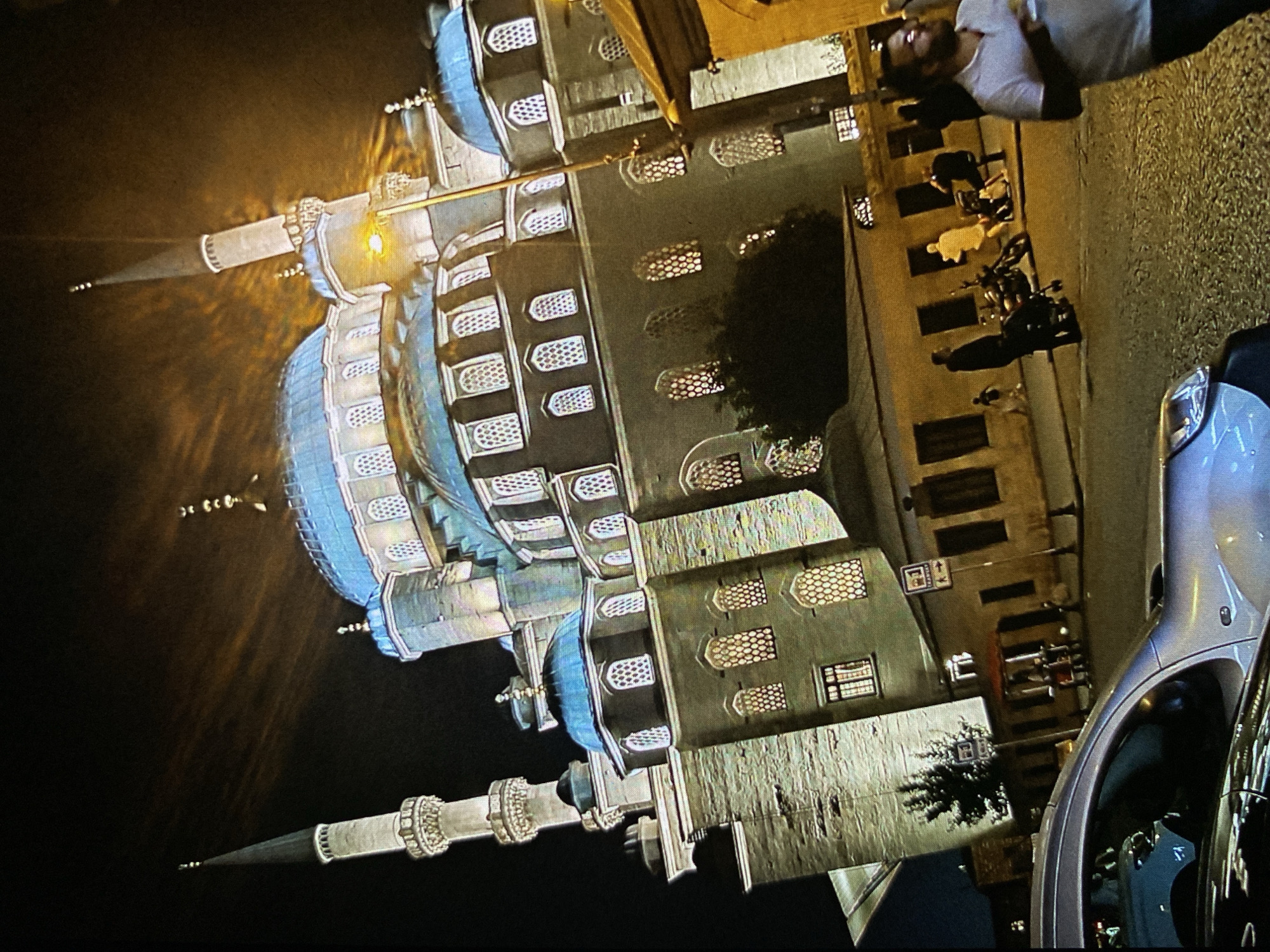 The image depicts a nocturnal urban scene dominated by a large, elaborately designed building reminiscent of Ottoman architecture, possibly a mosque, set against a dark sky. The structure features multiple slender minarets, some topped with spires and emitting a bright glow, and several prominent domes with a distinctive blue or teal hue. Its light-colored, textured facade is punctuated by numerous windows, many adorned with intricate grilles. Artificial lighting illuminates the building, with light spilling from some windows.
On the right side of the scene, a street or paved area is visible. One person, wearing a white t-shirt and dark pants, stands looking upwards towards the building. Further along the street, several motorcycles or scooters are parked, and other indistinct figures can be seen. The front section of a silver car is partially visible in the bottom right foreground. A small tree is present near the base of the main building. The overall ambiance is that of a city night, with dramatic lighting highlighting the architectural grandeur. There are a couple of small, illegible signs near the street. The location context suggests this scene is in Bankya, Bulgaria.