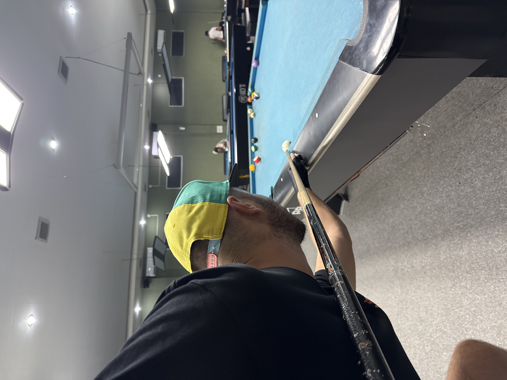 The image captures a close-up, low-angle view of a person playing billiards in Kuta Selatan, Indonesia. The person, seen from the back and side, is wearing a black t-shirt and a two-toned yellow and green baseball cap, facing towards a blue pool table. Their head is bent down, focused on aiming a cue stick, which is prominently featured in the foreground, extending towards the table. Several colorful billiard balls are scattered across the table. In the background, other pool tables are visible, with at least two other individuals playing or observing the game, also in Kuta Selatan. The environment appears to be an indoor establishment, with fluorescent lighting illuminating the scene and a muted green wall in the distance.