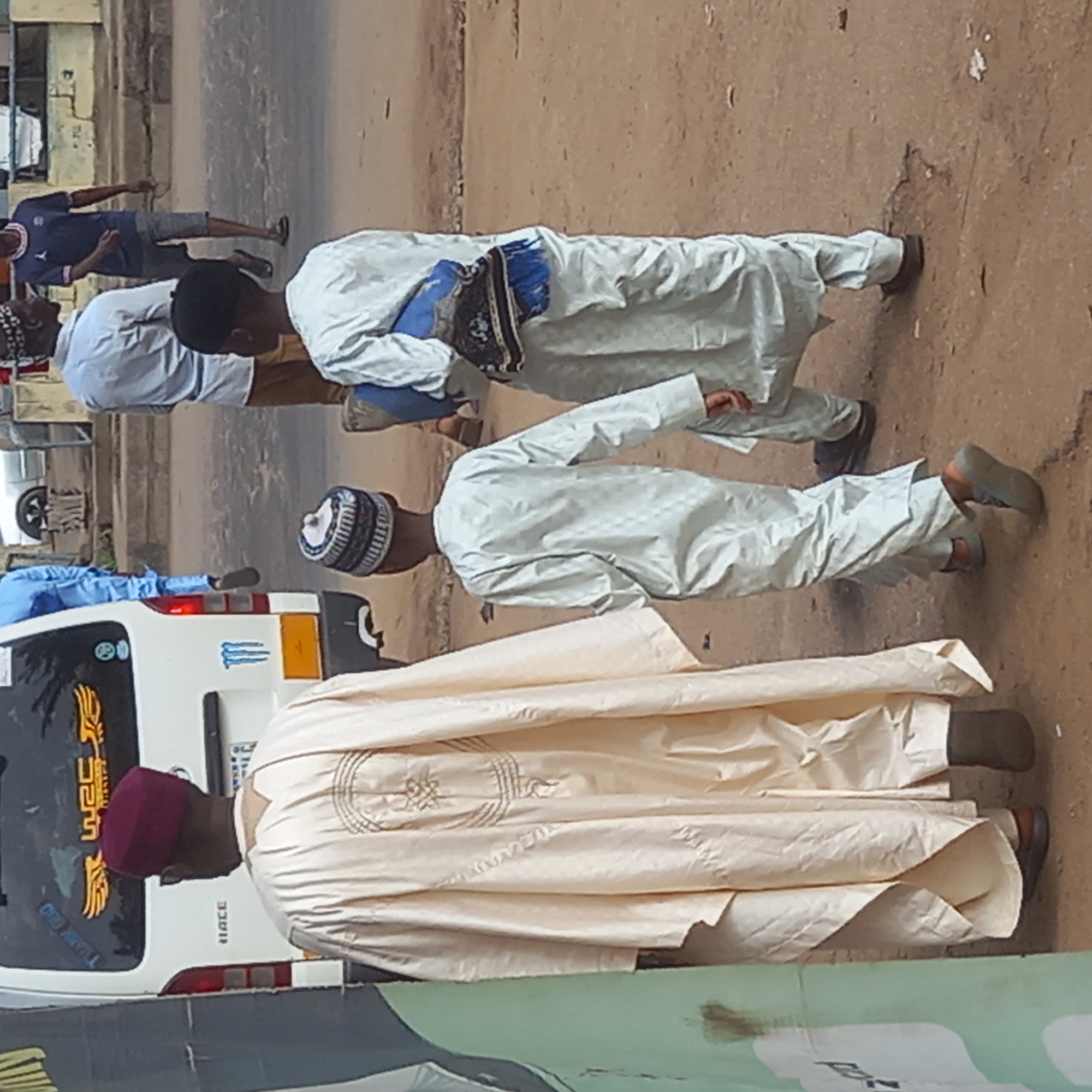 In the city of Dadi Riba, Nigeria, three men wearing traditional attire are walking along a dirt road next to a white van. The man closest to the camera wears a maroon cap and a long, light beige robe with embroidered circular designs. The middle man has a white and black cap and a white robe with textured patterns. The third man is wearing a white robe with a blue satchel. A few other people walk in the background, and the edge of a green poster is visible at the bottom of the frame.