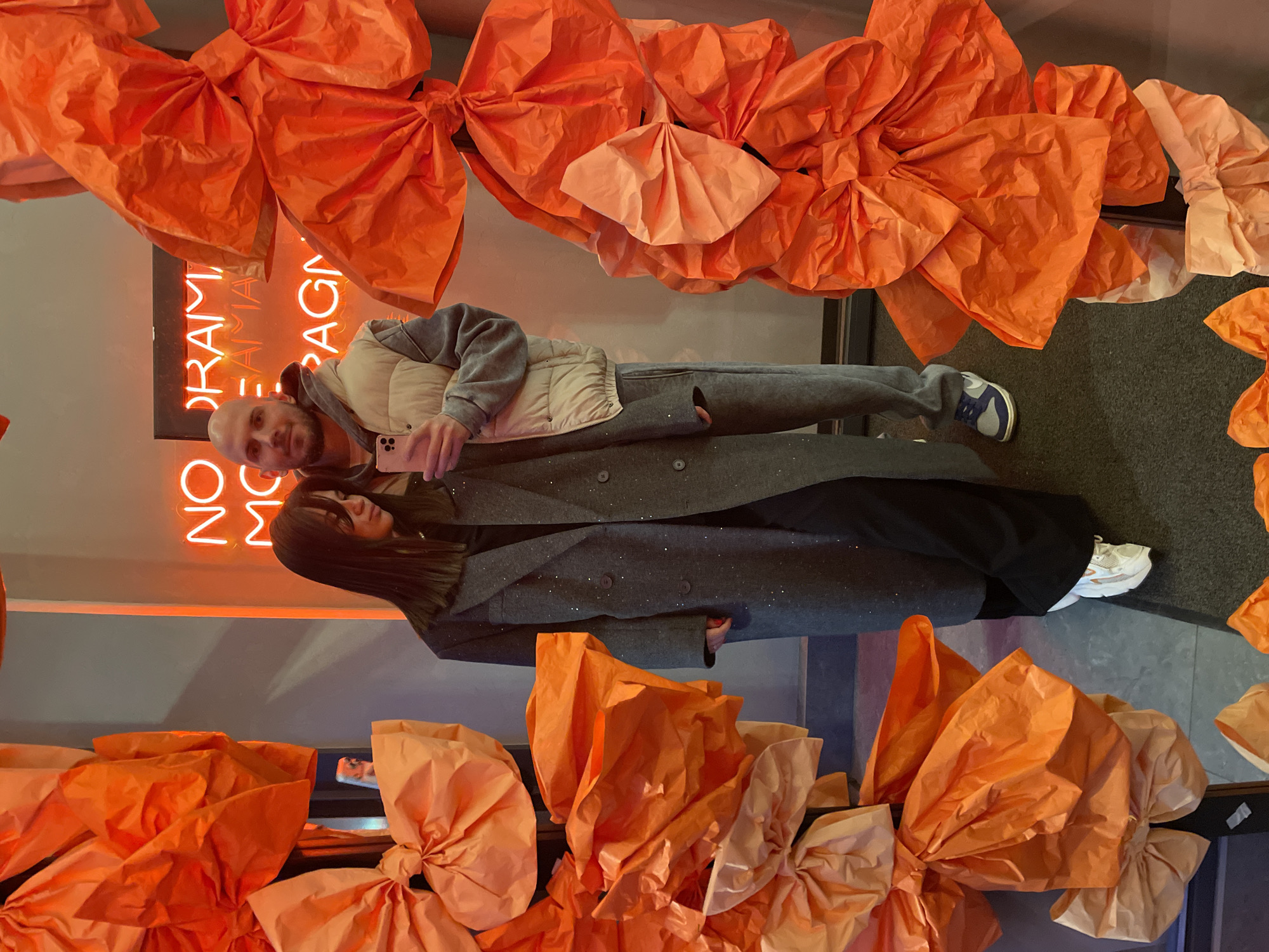 Two people, a man and a woman, are posing for a photograph in front of an orange-themed display. The location is likely in Krasnoyarsk, Russia. The man is bald, wearing a vest and grey sweatpants, and holding a phone up to take the picture. The woman has long brown hair and is wearing a grey coat and black pants, and white sneakers. The background is decorated with large orange paper bows. A neon sign partially visible reads "NO MORE DRAMA". The setting appears to be inside a building, possibly a store or event space.