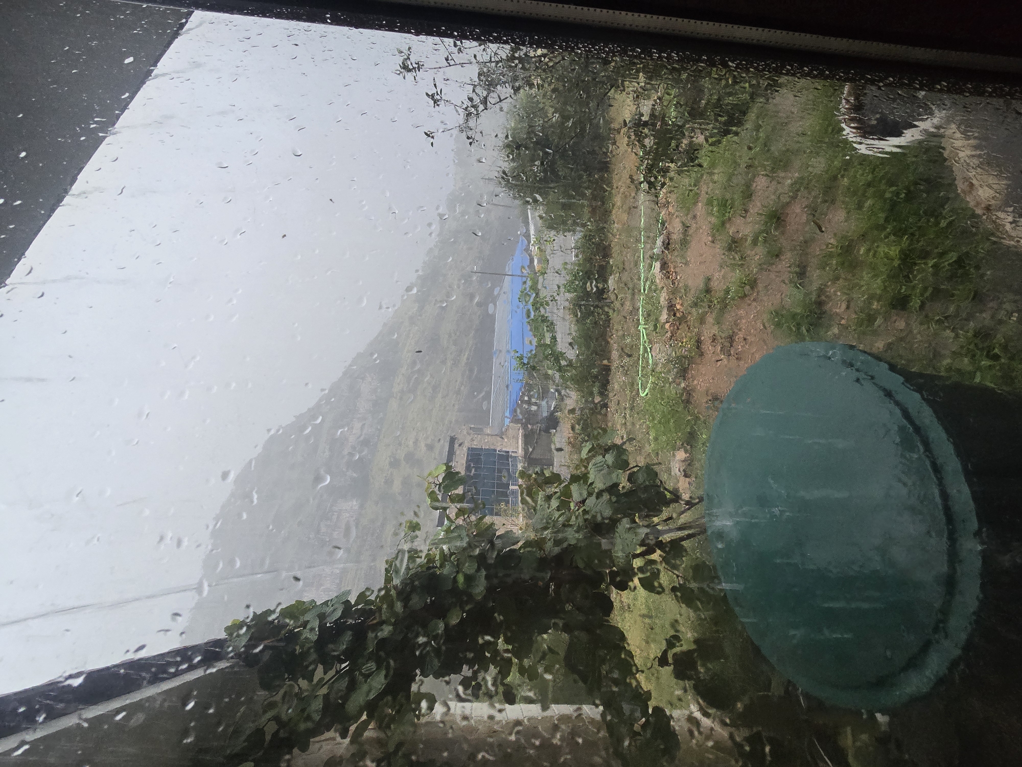 This image shows a view from a window during a rain shower, looking out onto an outdoor scene. The foreground features a large, dark green, circular lidded container, possibly a water tank. To the left, a building wall is partially visible, with a vine-covered structure arching over the scene. Beyond this, the ground is covered with sparse green vegetation and some exposed soil. A green hose lies on the ground. In the middle ground, there is a building with a blue tarp or roof in front of it, and a road or pathway. The background is dominated by a large, overcast sky and a distant, misty mountain or hillside. The window through which the scene is viewed is covered in raindrops, distorting the view and creating streaks. The overall impression is one of a rainy day in a semi-rural or undeveloped area, with a focus on the natural elements and weathered structures.