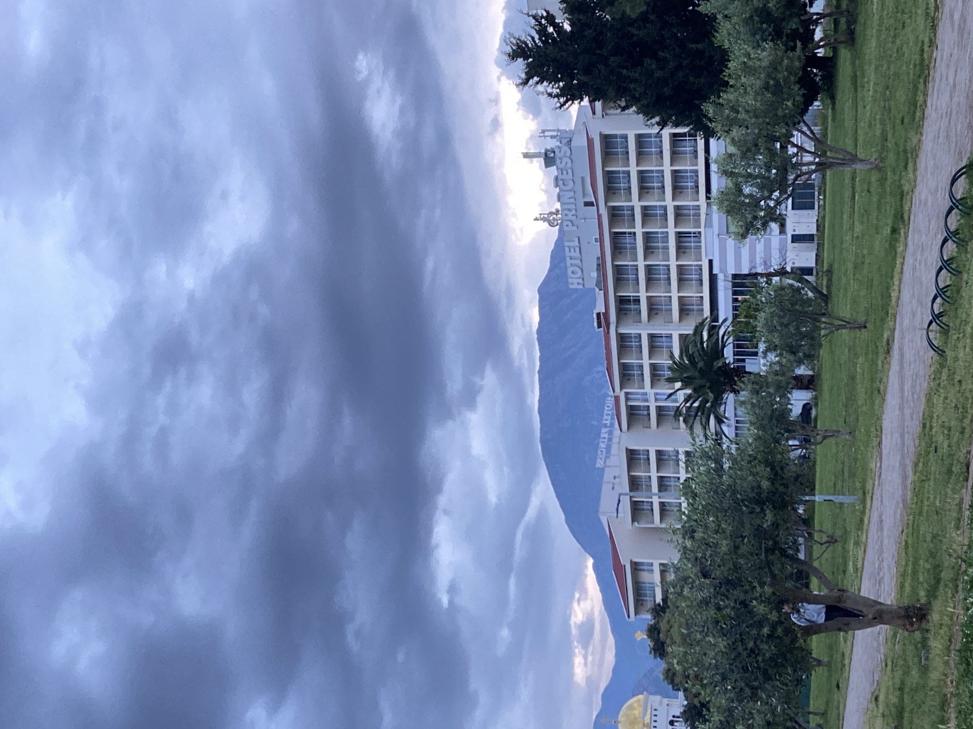 This landscape image captures a scene in Bar, Montenegro, centered around the "HOTEL PRINCESS." The hotel is a multi-story, light-colored building featuring many windows, with its name prominently displayed in white letters on the roof. Behind the hotel, a large, dark mountain range rises, its upper slopes partially obscured by dramatic, heavy grey and white clouds that dominate the sky. Some brighter patches of light break through the cloud cover, creating a somewhat diffused and moody atmosphere.
In the foreground, a well-maintained green lawn stretches out, dotted with various trees, including some taller, darker evergreens and several smaller trees with lighter green foliage. A paved pathway curves through the grassy area, and metal bike racks are visible alongside it on the right. In the lower left corner, partially hidden behind a tree, a person is visible wearing a light-colored top and dark pants. Further to the lower left, a distinctive golden dome and a portion of a white building, possibly a religious structure, can be seen. The overall impression is an overcast daytime scene, suggesting either an impending change in weather or simply a heavily clouded day.