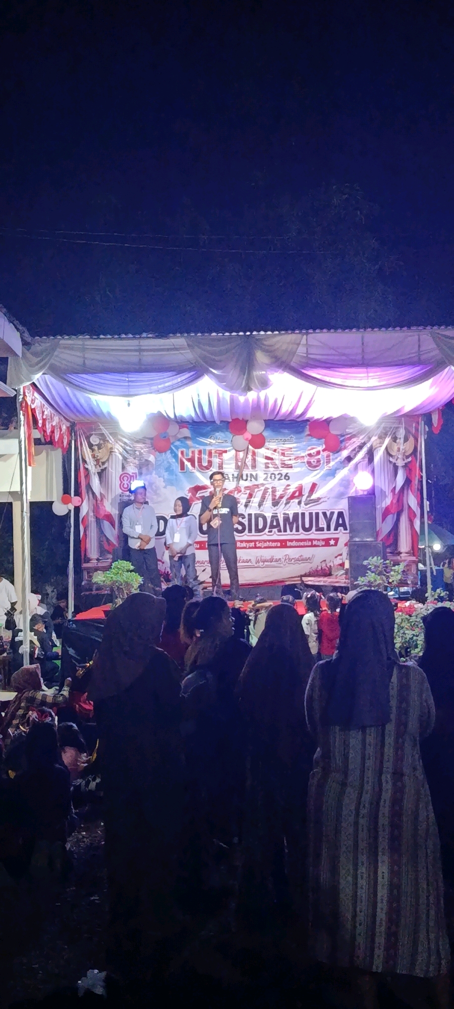 The image depicts an outdoor stage event at night, likely in Binangun, Indonesia, based on the banner text. A banner across the stage reads "NUTRI SE... FHUN 2026 FESTIVAL SIDAMULYA". Three individuals are on stage: a man in the center holding a microphone and speaking, a man to his left wearing a light-colored shirt and a blue cap, and a woman to the center-left wearing a light-colored shirt and a headscarf. The stage is decorated with red and white fabric draping and red and white balloons. The audience, mostly women wearing hijabs, is visible in the foreground, looking towards the stage. The environment is dark, indicating it is nighttime, with stage lighting illuminating the performers and the banner.