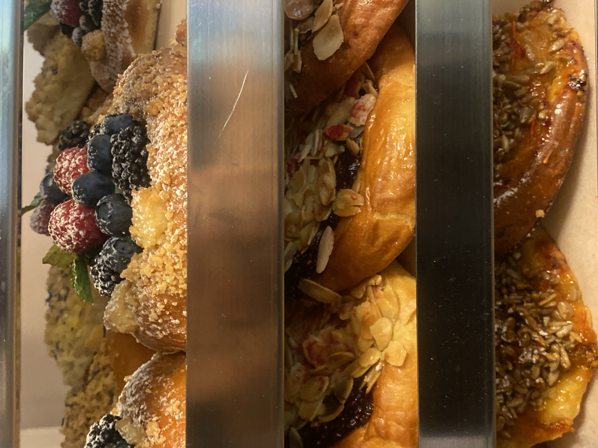 The image displays a close-up of various pastries arranged in what appears to be a display case. The pastries are positioned in vertical sections, separated by metal dividers.
On the left, a pastry is adorned with fresh berries, including raspberries, blueberries, and blackberries. It is also coated in a crumbly topping and dusted with powdered sugar. Next to it, a croissant-like pastry is filled with what looks like jam and topped with sliced almonds. Another pastry to the right is glazed and sprinkled with seeds, possibly sunflower or sesame seeds.
The pastries appear fresh and inviting, suggesting they are for sale. The lighting is bright, indicating an indoor setting, likely a bakery or pastry shop. There are no people visible in the image. The overall impression is one of a delicious assortment of baked goods. The context provided specifies the location as Warsaw, Poland.
