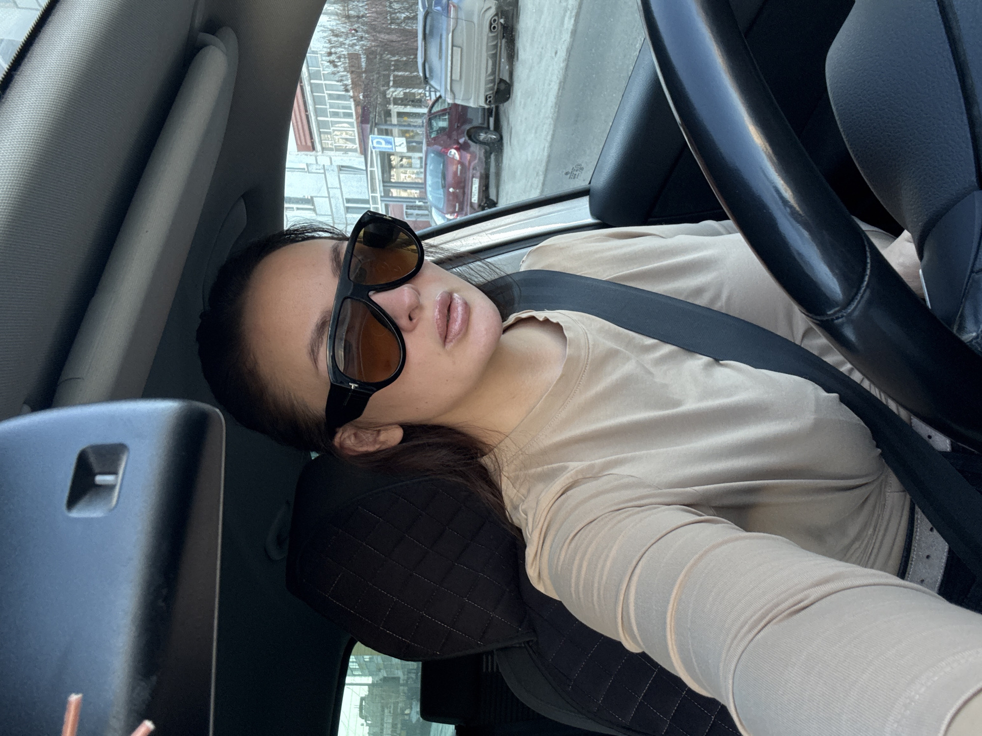 The image shows a woman sitting in the driver's seat of a car, wearing sunglasses and a seatbelt. The car is parked on a street in Chaikovsky, Russia, as evidenced by the cars and buildings visible through the window.