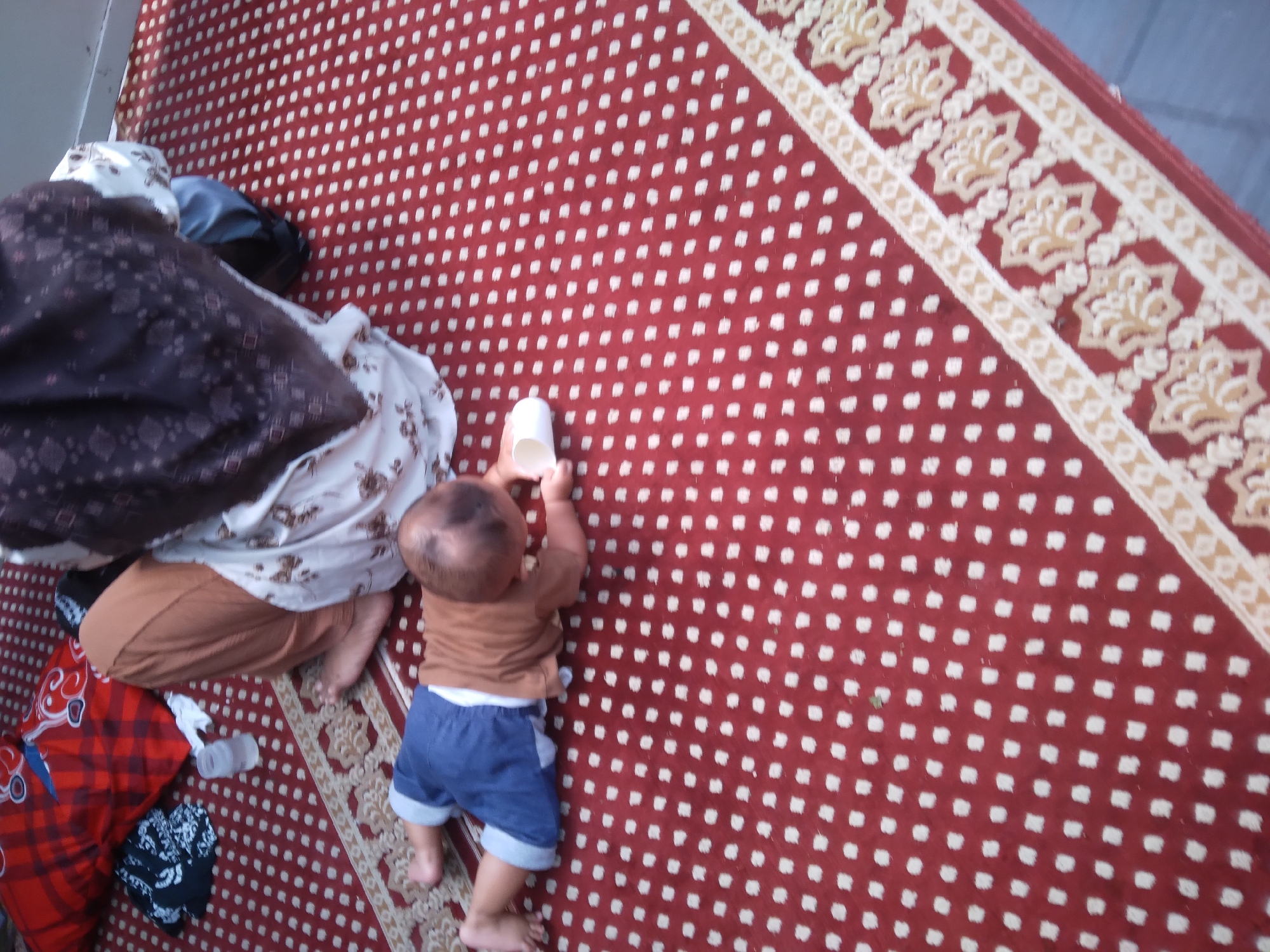 A baby is lying on a red carpet with a white dotted pattern in what appears to be a mosque. The baby is wearing a brown t-shirt and blue shorts. A woman in a brown hijab and a white and brown patterned dress is sitting behind the baby. She is holding a small white cup. There are also pillows and blankets in the background. The image is taken from a high angle, looking down at the baby. The carpet has a decorative border. The location is identified as Desa Pamijen Kecamatan Sokaraja, Indonesia.