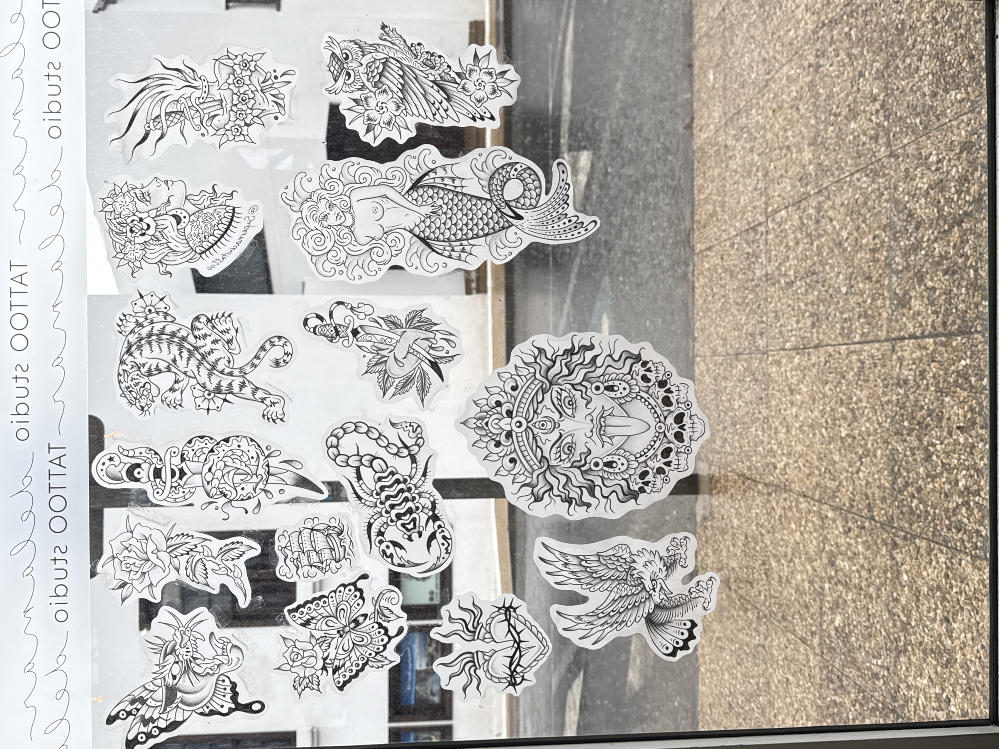 A sheet of black and white tattoo flash art is affixed to a window. The flash sheet is labeled "OOTATTOO OOTATTOO OOTATTOO." The tattoos displayed include a dagger impaling a bouquet of flowers, an owl, a mermaid, a panther, a dagger and snake, a scorpion, a sun/moon face with skulls, and an eagle. Through the window are glimpses of the external environment, including a grey street and an interior with books and other objects. The location is identified as Yaiza, Spain.