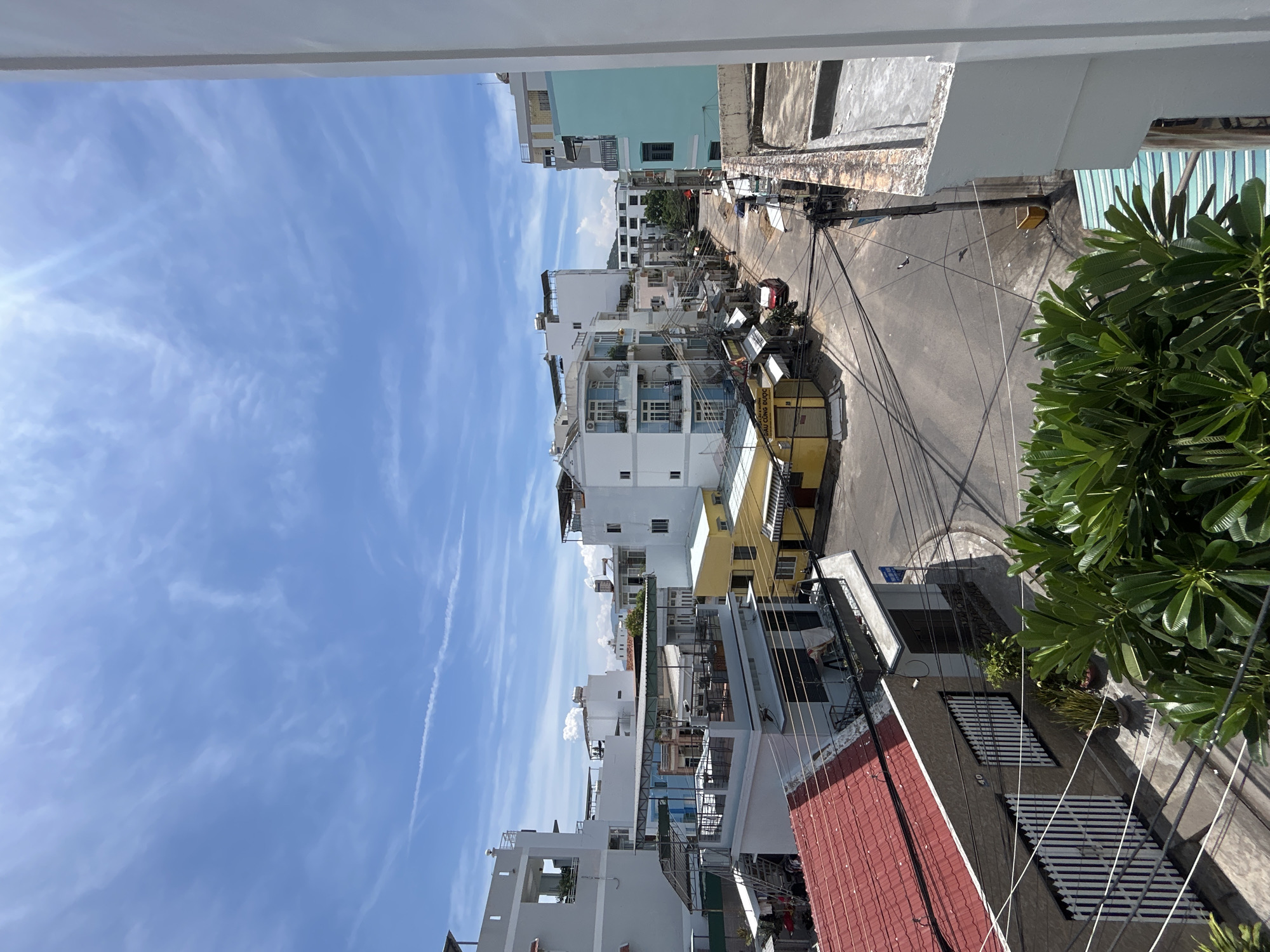 This image captures an aerial view of a street in Nha Trang Ward, Vietnam, under a bright, partly cloudy sky. The scene is characterized by a dense urban environment with a variety of multi-story buildings.
The foreground features lush green foliage, likely from trees or large shrubs, obscuring the lower portion of the frame. To the left, a building with a distinctive red tiled roof is visible, with white striped gates and fences. Above this, a light blue building with multiple balconies and windows is seen.
The street itself is paved and appears to be a typical urban road, with numerous power lines crisscrossing overhead, supported by utility poles. Several buildings line the street, displaying a mix of architectural styles and colors. Notable structures include a yellow building with awnings and a turquoise building on the right side of the street. Cars are present on the street, including a red car parked near the yellow building.
Further down the street, more buildings are visible, some with balconies and others appearing to be businesses. The sky is a vibrant blue with wispy white clouds and a bright sun casting strong light and shadows. The overall impression is a bustling, residential and commercial area in a coastal Vietnamese city.