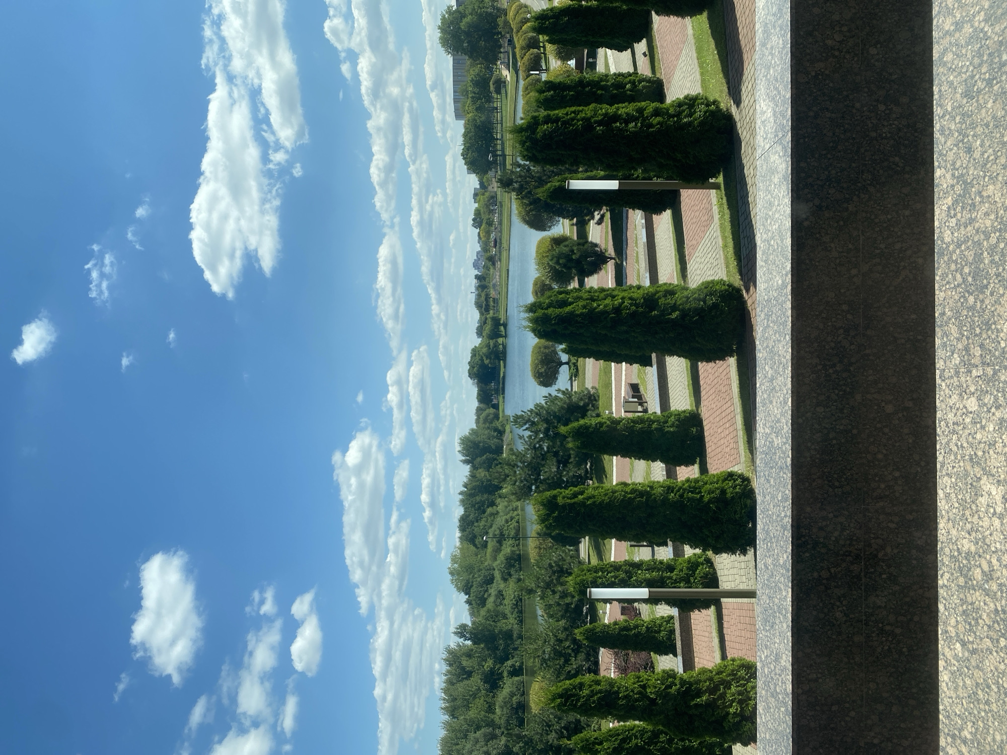 The image depicts a scenic view of a park in Minsk, Belarus, characterized by a serene lake under a bright blue sky dotted with white, fluffy clouds. In the foreground, a row of neatly trimmed evergreen trees stands tall along a paved walkway. The walkway is laid with interlocking bricks in a red and gray pattern, interspersed with patches of green grass. Beyond the trees and walkway, the tranquil lake stretches out, bordered by lush green foliage and trees that form a verdant backdrop. In the distance, several buildings can be seen, hinting at the urban surroundings of the park. The overall impression is one of a peaceful and well-maintained public space, likely a popular spot for relaxation and enjoying nature within the city.