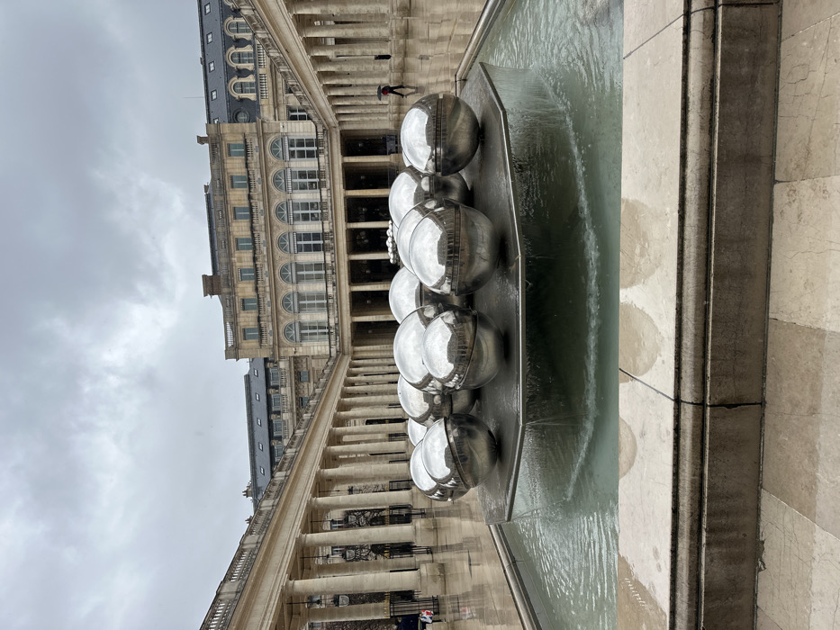 This image depicts the main courtyard of the Palais Royal in Paris, France, under an overcast sky. In the foreground, a modern art fountain occupies the center, featuring an arrangement of approximately 16 large, highly polished, spherical metallic objects clustered on a dark, octagonal platform. Water overflows from this platform into a wider, light-colored stone basin below, creating a ripple effect.
The courtyard is surrounded by the classical architecture of the Palais Royal, characterized by multi-story buildings with light-colored stone facades. The ground levels feature long arcades supported by rows of tall, round stone columns. The upper stories display numerous windows, some arched, and dark, sloped roofs with dormers. The paved ground of the courtyard appears wet and reflective, indicating recent precipitation.
Two individuals are visible: one figure walks on the far right side of the courtyard, away from the viewer, holding an open red umbrella. A second, partially obscured figure is present on the far left side of the courtyard near the columns. The sky is uniformly grey and cloudy.