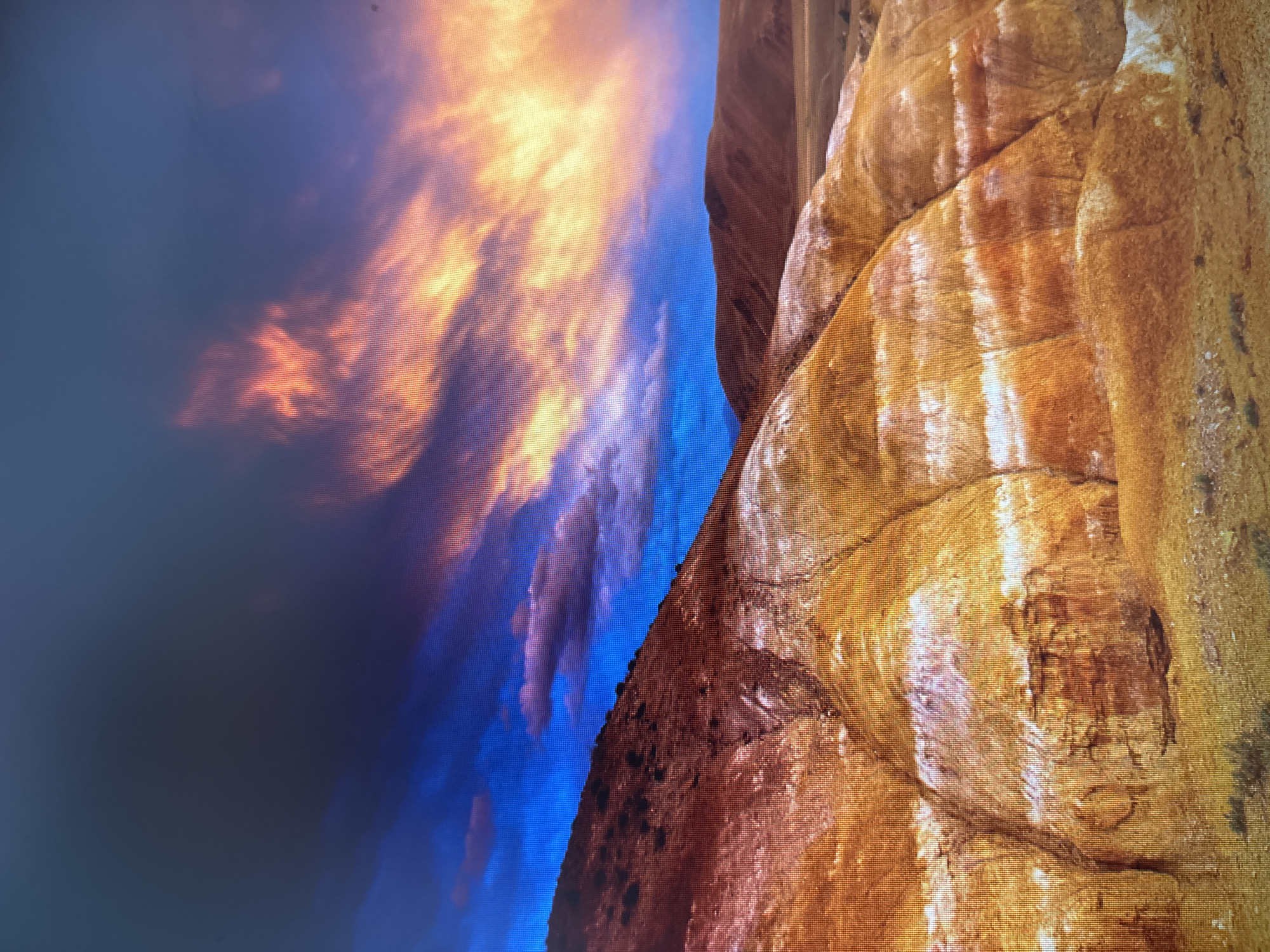 The image depicts a landscape with prominent geological formations. On the right, there's a striated rock formation with bands of brown, tan, and white. The rock appears textured and possibly sedimentary. To the left of the rock is a darker, possibly mountainous, ridge. Beyond that, the image shows a vivid sky with shades of blue, purple, and orange, resembling either a dramatic sunset or sunrise. The image does not depict any people, specific actions, or interaction. There's nothing in the image to suggest that it is located in Saint Petersburg, Russia.
