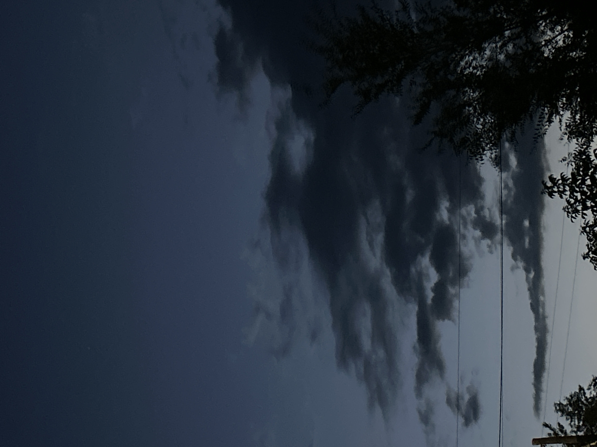 The image depicts a dusky sky with dark, textured clouds. The sky transitions from a deep blue at the top to a lighter shade closer to the horizon, suggesting twilight or a pre-storm atmosphere. Silhouetted against the sky are the branches of trees on the right side, and what appear to be power lines stretched horizontally across the middle portion of the frame. A slender pole, possibly a utility pole, is partially visible on the far left. The overall scene is dark and somewhat dramatic, with an emphasis on atmospheric conditions and man-made infrastructure. The location, Майқайың кенттік әкімдігі, Kazakhstan, implies an outdoor setting, likely on the outskirts of a settlement or in a more rural area given the presence of trees and utility lines.