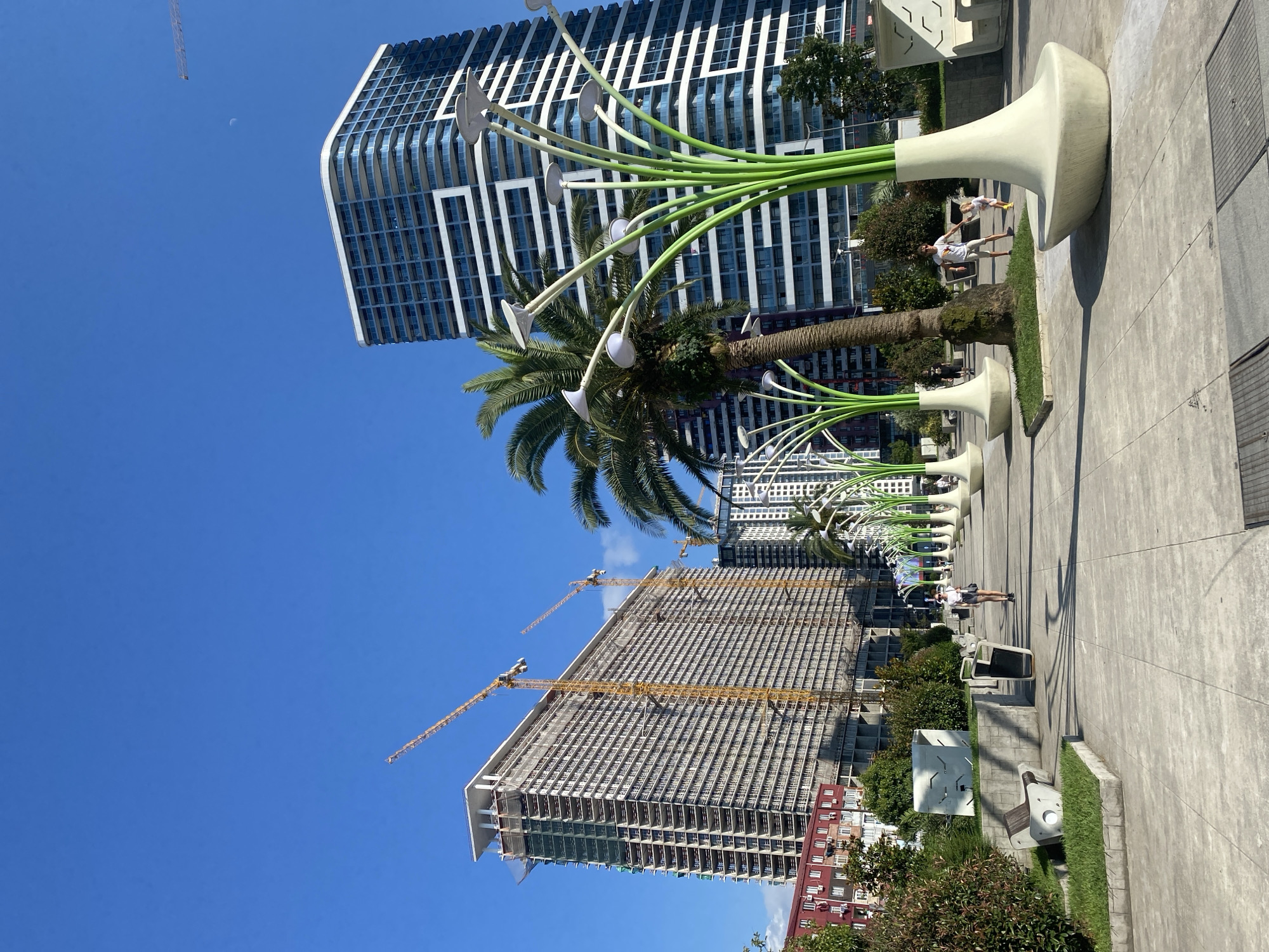 This is a medium-shot, eye-level, outdoor photograph taken in Batumi, Georgia, likely on the Batumi Boulevard. The scene captures a sunny day with a clear blue sky. In the foreground, a paved walkway is lined with distinctive, modern, light-colored planters that contain tall, green, sculptural elements resembling stylized flowers or plants with white flower-like lights at the top. A palm tree is visible to the left of the walkway, behind some of these planters.
Further down the walkway, several people are walking. To the right, a man in a white t-shirt and shorts is holding the hand of a young child in a yellow outfit. To the left, a person in a white top and dark shorts is walking away from the camera.
In the middle ground and background, there are several large buildings. On the left, a massive, multi-story building is under construction, with several yellow construction cranes visible against the sky. This building appears to be clad in scaffolding and netting. To the right of the construction site, another, completed, modern building with a dark facade and many horizontal balconies is visible. Dominating the right side of the background is a tall, sleek, modern skyscraper with a white and dark blue facade and numerous balconies.
The environment appears to be a public space designed for leisure and recreation, with the modern architecture contrasting with the natural elements of palm trees and greenery. The presence of construction indicates ongoing development in the area.