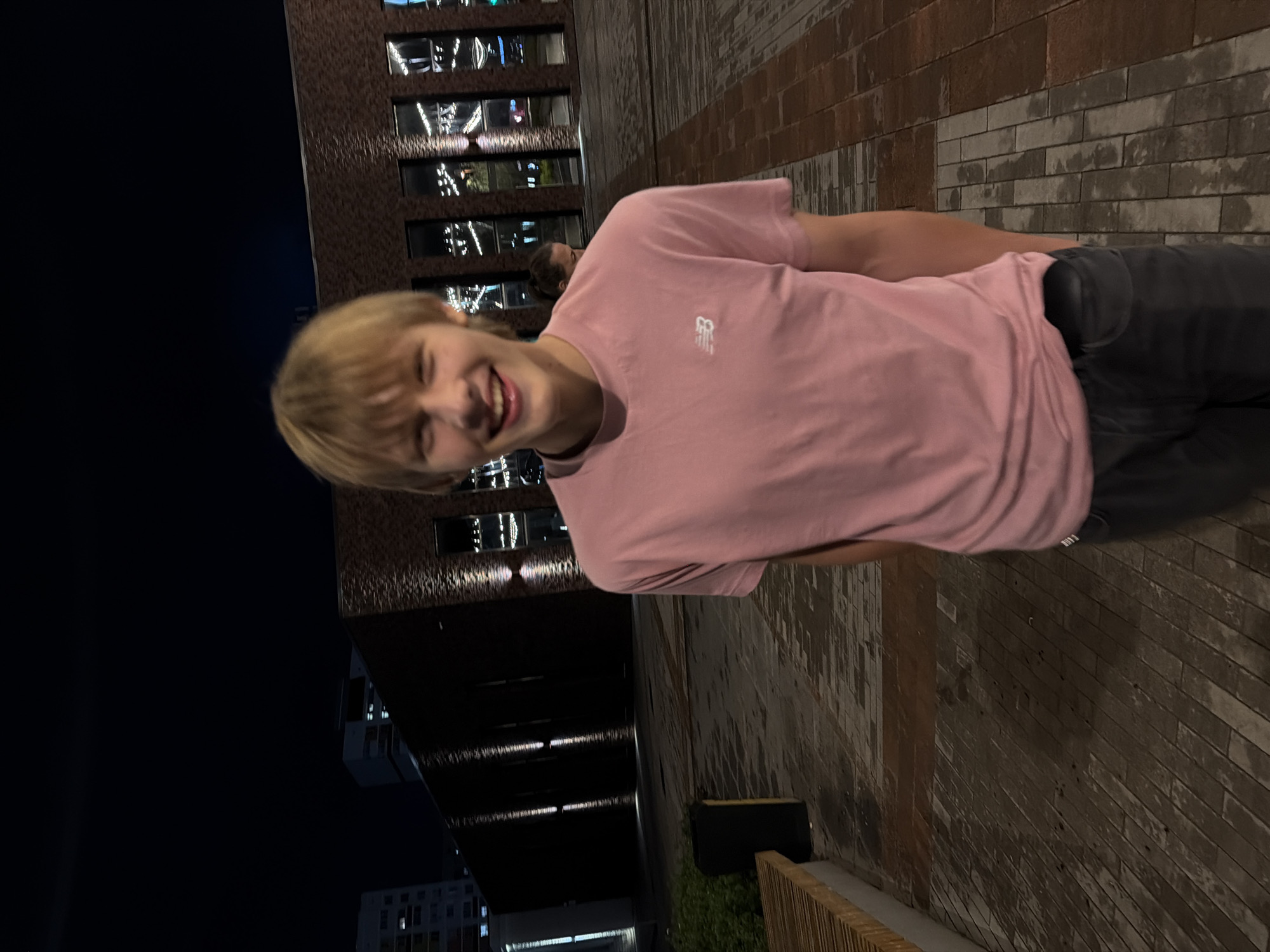 A young male with blonde hair and a pink t-shirt with the New Balance logo is smiling at the camera at night. He is standing on a brick-paved walkway in front of a modern building with large windows and lights. The building appears to be a commercial or public space. There is a bench to the left of the frame, and a trash receptacle behind it. The sky is dark, indicating it is nighttime. The location is identified as Izhevsk, Russia.
