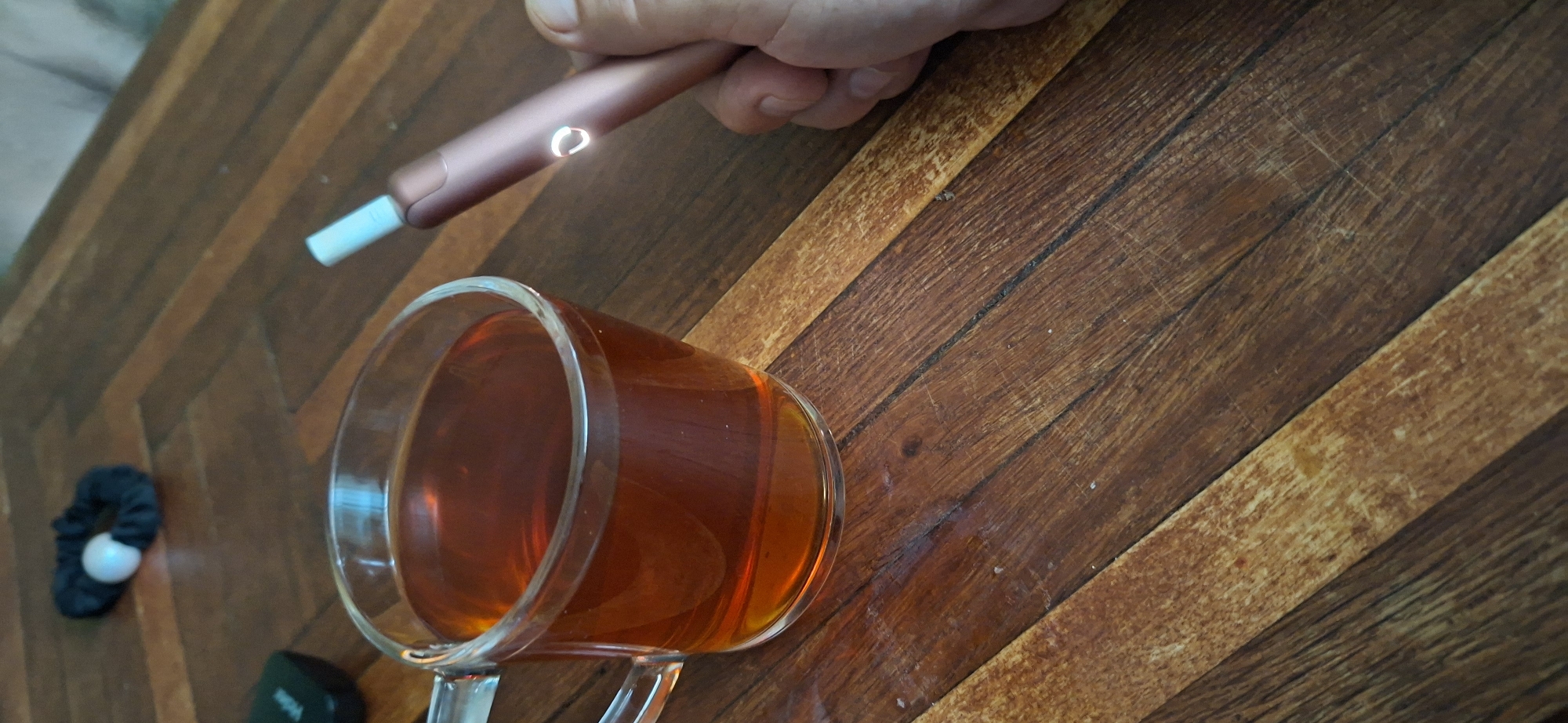 A glass mug filled with amber-colored liquid, likely tea, sits on a wooden table. To the right of the mug, a hand holds a rose gold colored electronic cigarette device, which is lit, emitting a faint glow from its indicator light. In the background, a black hair scrunchie with a white pearl-like accent lies on the table. The table surface exhibits a wood-grain pattern, with distinct planks arranged diagonally. The scene appears to be indoors, possibly in a home in Shakhtarsk, Ukraine.