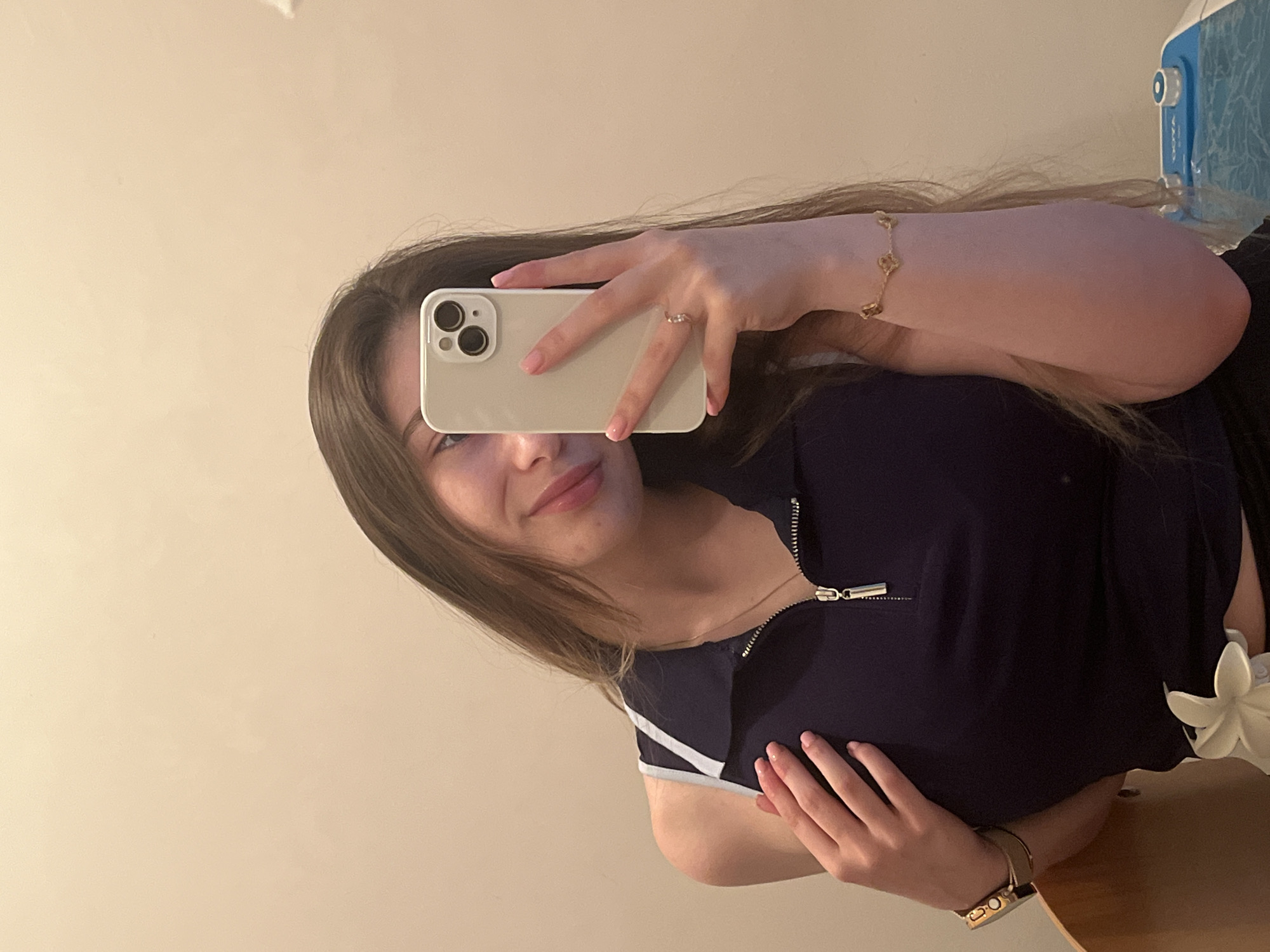 A young woman is captured in a mirror selfie, taken indoors in what appears to be a home setting. She is holding a light-colored iPhone, which partially obscures her face, though her lips show a slight smile. Her long, straight brown hair falls over her shoulders.
On her right wrist, she wears a gold bracelet with clover-shaped charms, and a ring is visible on her right ring finger. She is dressed in a dark blue top featuring a silver zipper at the neckline. A white strap, possibly from a bra or another layer of clothing, is visible on her left shoulder. Her left hand rests on her midsection, showcasing a watch with a brown strap on her left wrist.
To the lower right of the image, a white floral accessory is visible. The background consists of a plain, light-colored wall. On the far right, a blue and white appliance, possibly a humidifier or air purifier, with the text "OQYA" faintly visible on its side, can be seen. A light brown, possibly wooden, surface is present in the bottom right corner of the frame. The overall atmosphere is casual and personal.