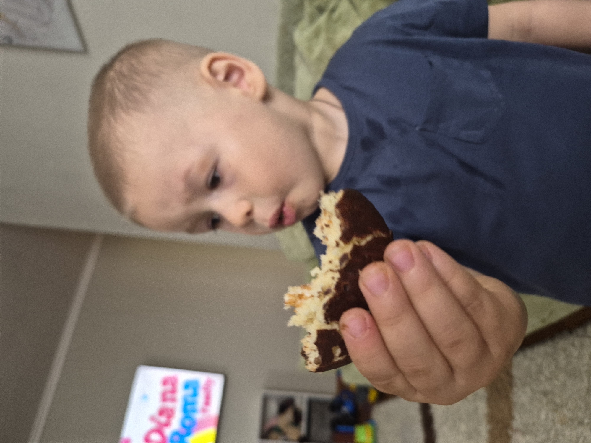 A young child with short blond hair, wearing a blue t-shirt, is holding a partially eaten chocolate-covered cake in their right hand. The child is looking at the cake with their mouth slightly open, appearing ready to take another bite. In the background, a television screen displays a colorful image, and part of a room with a wall, a rug, and other furniture is visible. While there is no explicit location information in the image, the caption mentions Tobolsk, Russia, so it is inferred the image was taken there.