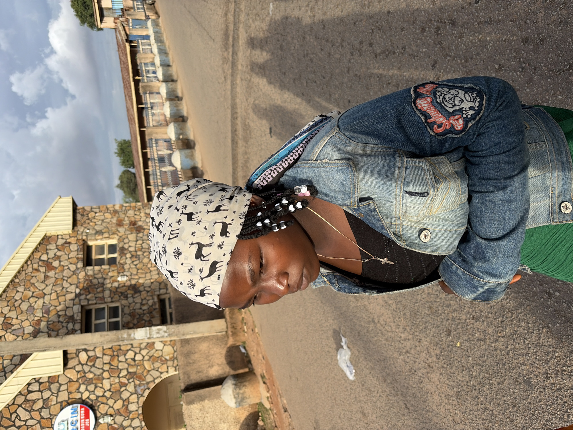 An outdoor, eye-level, medium shot depicts one young person standing on a paved road in Jos, Nigeria, under a partially cloudy sky. The subject, appearing to be female with dark skin, stands with her body slightly angled to the right, facing downwards and to her left (viewer's left), exhibiting a serious or pensive expression.
She wears a light-colored headwrap patterned with dark deer silhouettes, covering her hair which is styled in braids adorned with white and pink beads. A light blue denim jacket is worn over a black top with subtle embellishments, and a green, textured garment is visible below the jacket. A thin gold-colored necklace with a small silver-colored crucifix pendant hangs around her neck. On her right jacket sleeve (viewer's left), there is an embroidered patch featuring a white, cartoonish dog-like character and text in red and white that appears to read "SCHMOLTY NOO" or similar stylized words.
In the background to the left, a building constructed of irregular brown and tan stones features a light yellow corrugated metal roofline, an arched entrance, and two rectangular windows. A round sign, clearly visible on this building, reads "CINCIN NADIN JEWELRY JOS". A grey utility pole stands partially in front of this building. Further down the road to the right, another long, low building with a reddish-brown corrugated roof is visible. A low wall with blue-painted metal fencing and blue-and-white concrete bollards or barriers lines the road in front of this building. The asphalt-like road surface is dry, with a small piece of white plastic litter near the subject's feet on the left. Long shadows of two or more individuals, including one clearly holding a camera or phone, are cast on the road to the right, indicating low sun angle, consistent with early morning or late afternoon lighting. Sparse green trees are visible in the distant background.