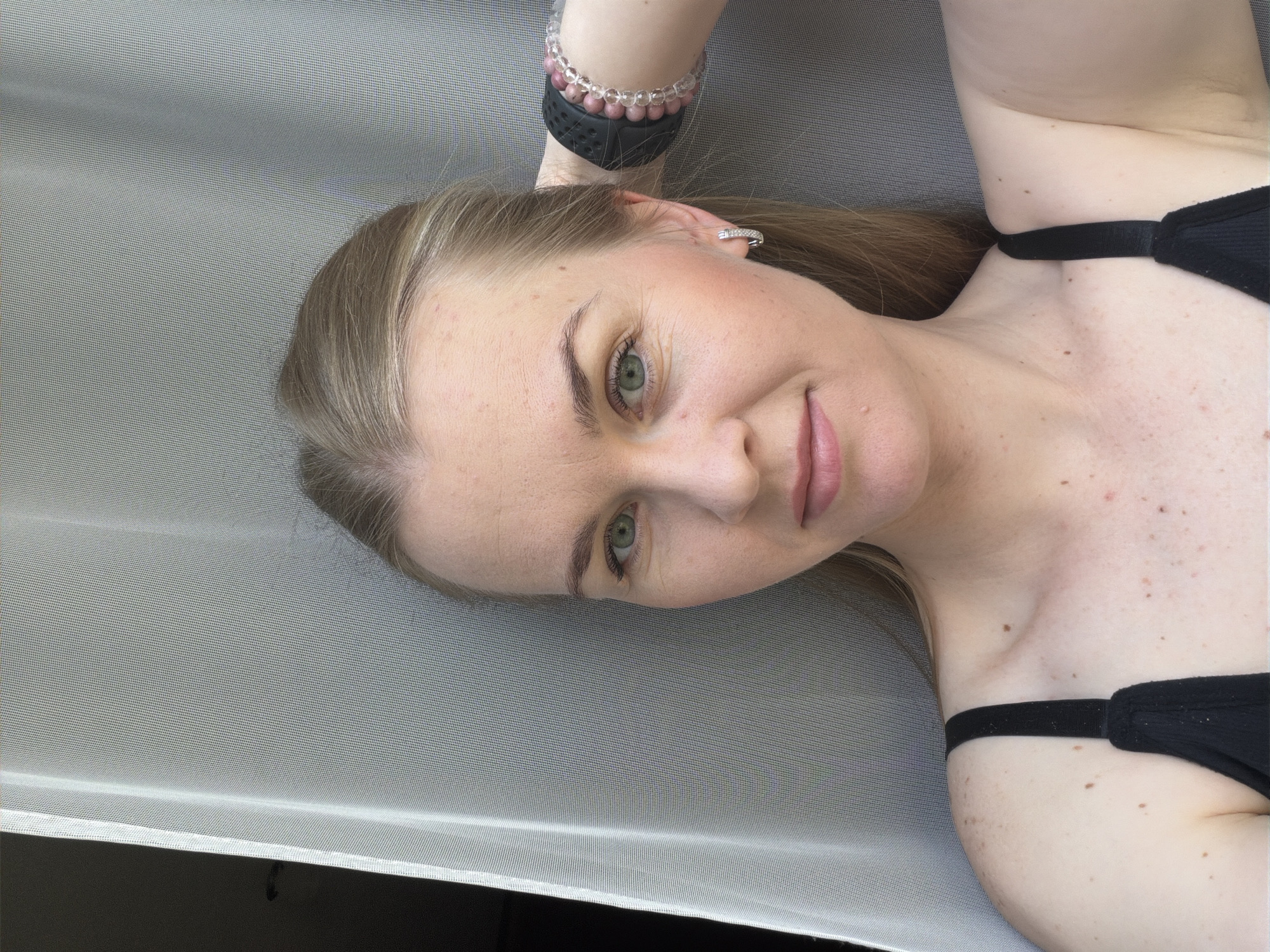 A young woman with fair skin, blonde hair, and green eyes is pictured from the chest up. She is looking directly at the camera with a slight smile. Her hair is pulled back, and she has a silver hoop earring in her left ear. She is wearing a black camisole and bracelets on her right wrist. The background consists of a textured grey curtain and a dark, out-of-focus area. The lighting suggests an indoor setting, possibly near a window. The specific location in Khabarovsk, Russia, cannot be determined from the image alone.