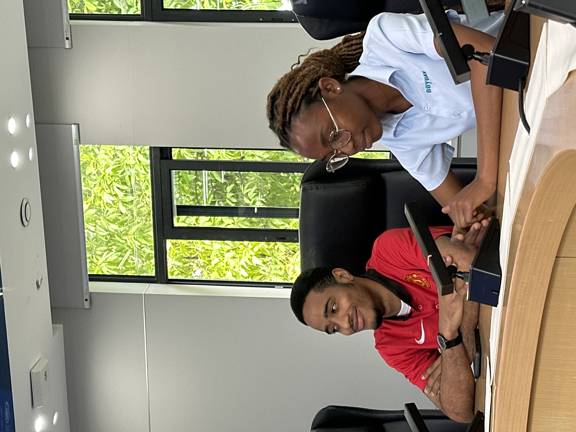 The image, set in Jiangjin, China, captures two young adults engaged at a workstation within a modern indoor environment. Bright natural light streams through large windows, revealing lush green foliage outside, indicating it is daytime.
On the left, a young woman with dark skin and braided hair wears thin gold-rimmed glasses and a light blue collared shirt with the word "BOYUAN" printed on it. She leans forward, her gaze directed downwards, seemingly interacting with an electronic device or screen on a black stand positioned on the light wooden desk in front of her.
Beside her, on the right, a young man with dark skin and short dark hair also looks down with a slight smile. He is wearing a red collared shirt with a white Nike logo and a club crest, likely Manchester United. A watch is visible on his left wrist, and a mobile phone rests near his hands. Both individuals are seated in black office chairs, seemingly focused on their respective tasks on devices also mounted on black stands.
The room features light grey walls and recessed lighting fixtures on the left, contributing to a clean and functional atmosphere. Black frames border the windows, contrasting with the vibrant green outdoor scenery. The scene suggests a collaborative or individual work session, possibly in an office, classroom, or study area.