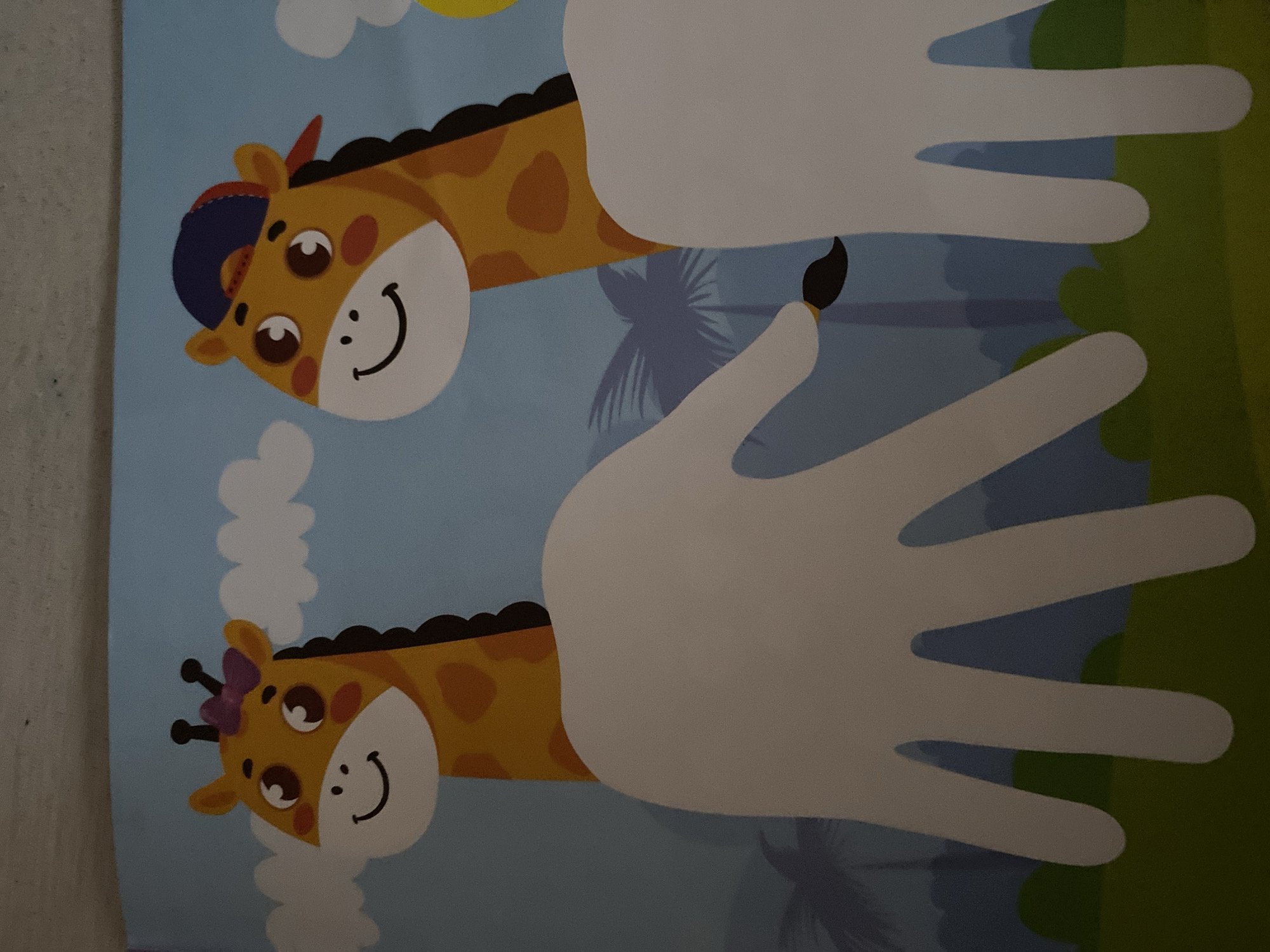 The image depicts a cartoon illustration of two giraffes with large, friendly eyes and smiling mouths against a backdrop of a pale blue sky with white clouds and silhouetted palm trees. The giraffe on the left wears a purple bow on its head, while the giraffe on the right sports a blue and red baseball cap. In the foreground, two large, white, hand-shaped cutouts are positioned in front of the giraffes, with the left cutout partially obscuring the left giraffe's body and the right cutout partially obscuring the right giraffe's body. The lower portion of the image shows a green, grassy landscape. The image is flat, with no discernible depth or dimensionality beyond the layered cartoon elements. There are no people, animals, or objects from Zgrade, Montenegro visible in this image.