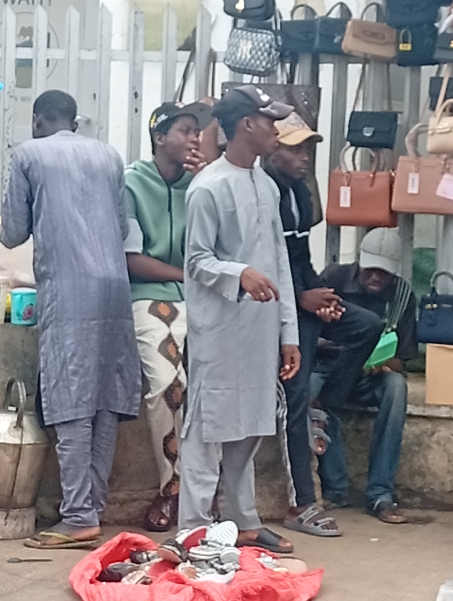 The image depicts a street scene in Maiburiji, Nigeria. Five men are visible, standing or sitting near a sidewalk and fence displaying handbags. The man on the left is wearing a long, gray traditional garment. Next to him, a man in a green hoodie and patterned trousers stands. A third man wears a gray shirt and pants and a baseball cap. Another man stands in a black jacket. On the right, a seated man is wearing a baseball cap and jeans. A red bag full of shoes is on the ground in front of them. Various handbags are displayed on the fence behind the men.