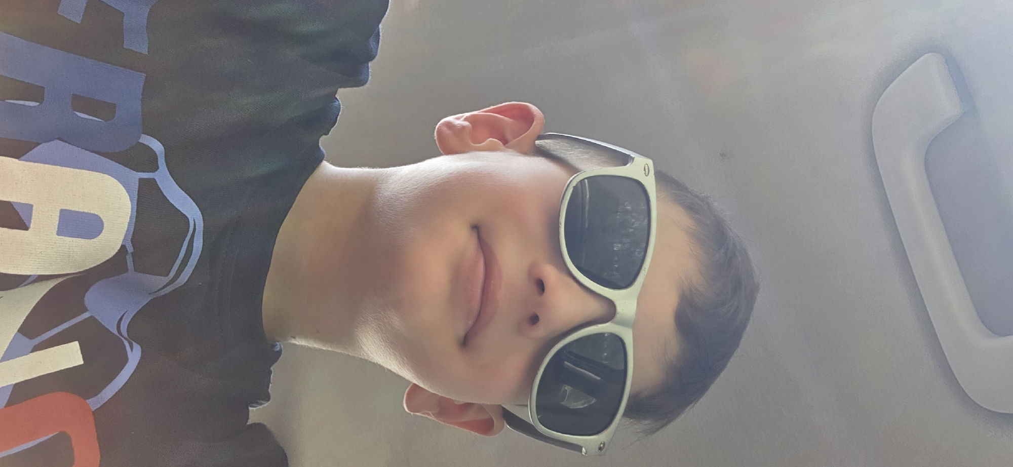 A young boy wearing sunglasses and a dark t-shirt is pictured from a low angle in what appears to be the interior of a vehicle. The boy has brown hair and is looking upwards towards the camera. The sunglasses have light-colored frames and dark lenses, reflecting the surroundings. The t-shirt has a graphic design on it with blue and white lettering. The ceiling of the vehicle, which includes a grab handle, is visible in the background.