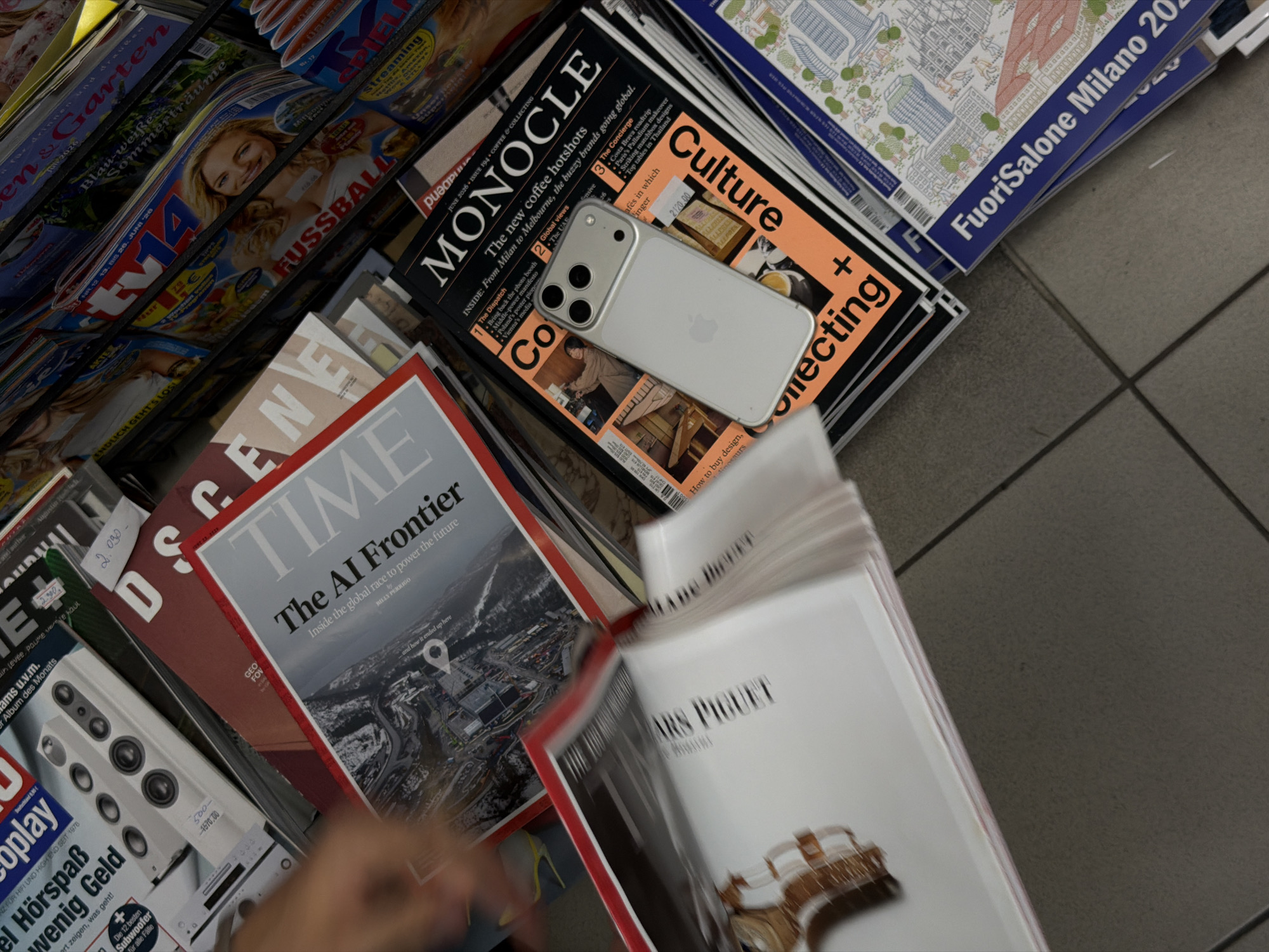 The image shows a close-up, overhead view of a display of various magazines and publications, likely in a store or newsstand. The scene is cluttered, with magazines stacked and fanned out, creating a sense of abundance and variety. Dominating the foreground is an iPhone placed on top of a magazine titled "MONOCLE." The iPhone is silver and features a triple-lens camera system.
The "MONOCLE" magazine has an orange and black cover with the headline "Culture + Collecting." Below this, smaller text reads "The new coffee hotshots" and lists articles like "The Dispatch," "The Concierge," and "Global views." Other visible magazine titles include "TIME," with the cover story "The AI Frontier," and "FUSSBALL" featuring a smiling blonde woman. There are also magazines with titles in German, such as "Garten" and "Streaming." Further to the right, a publication with colorful illustrations and the text "FuoriSalone Milano 2023" is visible.
In the lower left of the frame, a person's hand and arm are partially visible, suggesting they are browsing or interacting with the display. The lighting appears to be artificial, consistent with an indoor setting. The overall impression is one of a busy retail environment where a diverse range of reading material is available. The presence of both international and local publications hints at a cosmopolitan setting. The image is sharp in the foreground, with some motion blur on the magazines further back, indicating a slight movement of the camera or the subjects.