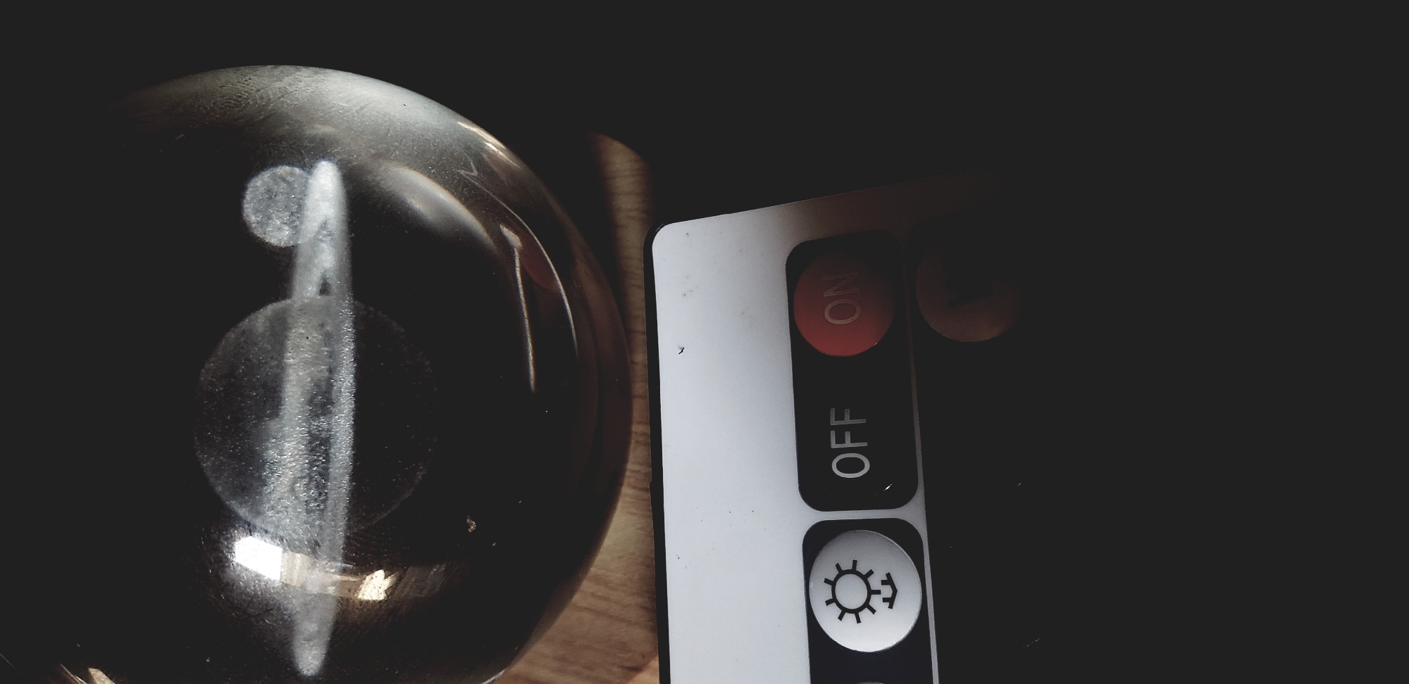 The image depicts a dimly lit scene with two primary objects: a glass sphere and a remote control. The glass sphere, located on the left side of the frame, appears to be resting on a wooden surface. It has a reflective surface, capturing highlights and creating a somewhat distorted reflection within its form. To the right of the sphere is a white remote control with black buttons and red "ON" lettering. The buttons are labeled "ON," "OFF," and feature a sun-like symbol. The background is dark and indistinct. The location is not evident from the image.