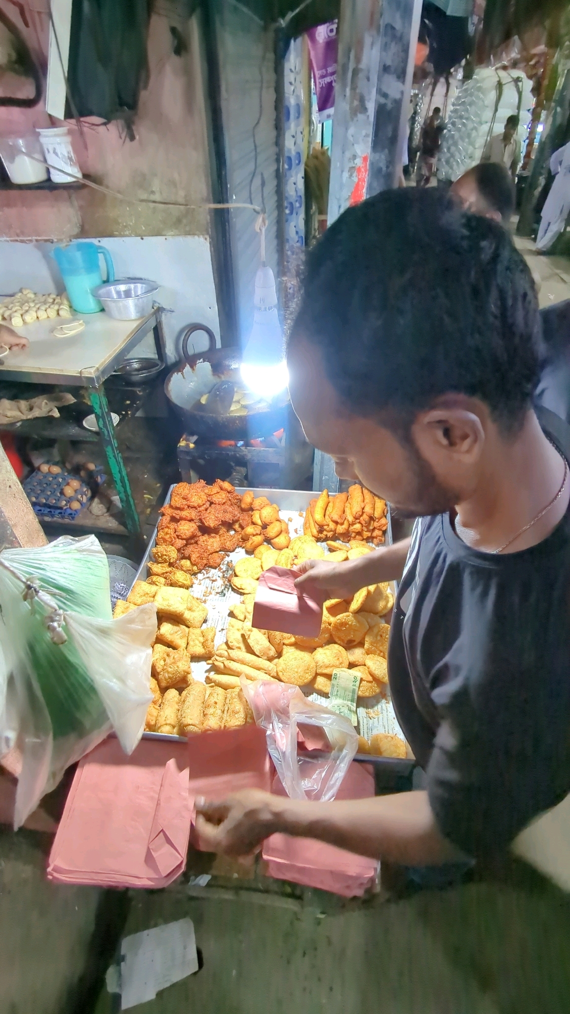 A man, seen from behind and in profile, wearing a black t-shirt, stands at a food stall in Dhaka, Bangladesh. He is holding a pink paper packet and appears to be in the process of serving food. In front of him, on a metal tray, is a large assortment of fried snacks, including what appear to be pakoras and other fried dough items. To the left, there are stacks of pink paper packets. Behind the man, cooking equipment, including a large wok over a flame, is visible. The background suggests a crowded market environment with other people and stalls. The lighting is dim, with a bright light bulb illuminating the food display.