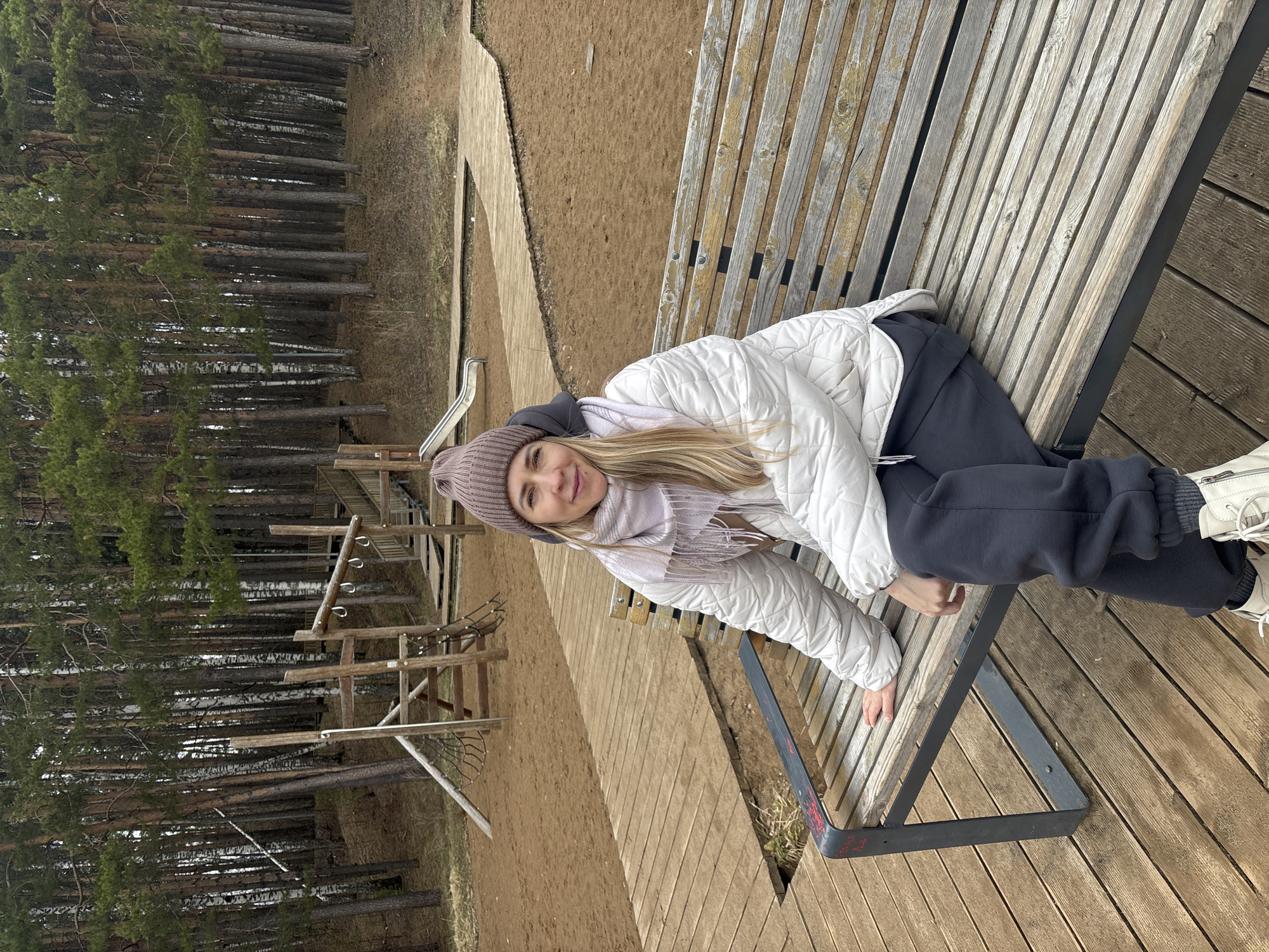 A woman is lying on a wooden bench in what appears to be a public park in Chaikovsky, Russia. She is wearing a mauve hat, a white quilted jacket, a light-colored scarf, and dark pants. Her blonde hair is visible. Behind her is a playground structure made of wood and a forest of tall, thin trees. The ground is covered with sand and wood planks.