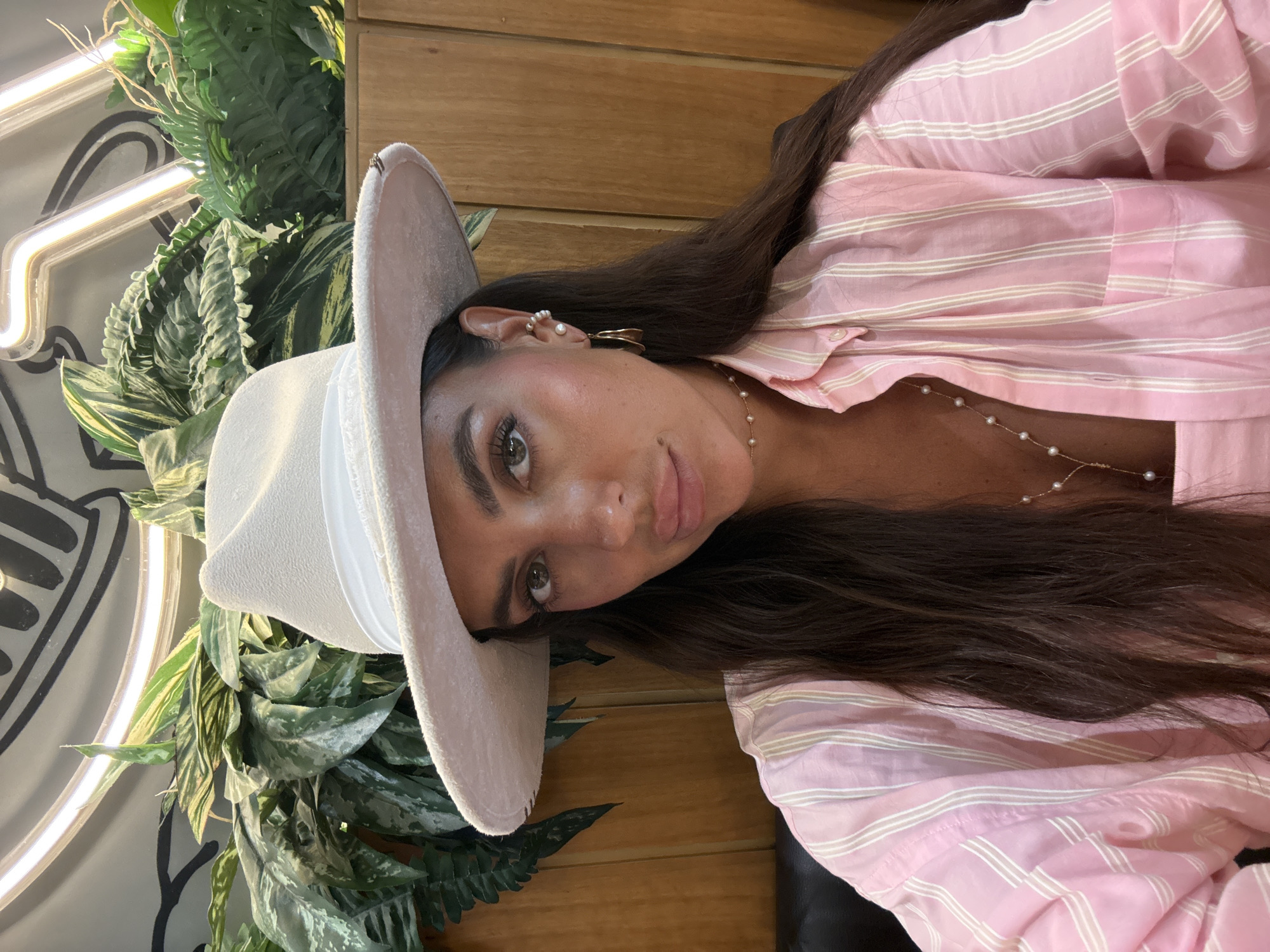 A close-up, waist-up selfie features a woman wearing a white fedora with a white band and a pink and white striped button-up shirt. She has long, dark hair and is adorned with gold hoop earrings and a pearl necklace. The background shows lush green plants, a wooden wall, and a neon light fixture. The scene appears to be set indoors, possibly in a restaurant or cafe, with the warm lighting and natural elements suggesting a relaxed, contemporary atmosphere. There is no clear indication of the specific location within Dubai, United Arab Emirates.