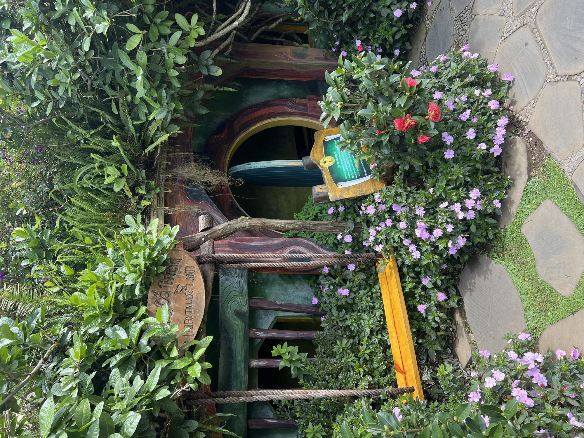 The image shows a whimsical, hobbit-hole-like structure nestled amongst lush greenery. A wooden sign above the entrance reads "Fairytale Land" with some Vietnamese text above it, likely indicating the location is a themed attraction. A yellow swing hangs in front of the entrance, its ropes attached to wooden beams. The structure itself has a rounded doorway and is painted in earthy tones with whimsical details. Pink and red flowers bloom profusely around the base of the structure and along the stone pathway leading to it. The environment appears to be an outdoor garden or park setting, possibly in a tourist area given the signage and design. This scene is located at the **Da Lat Fairytale Land** in Da Lat, Vietnam.