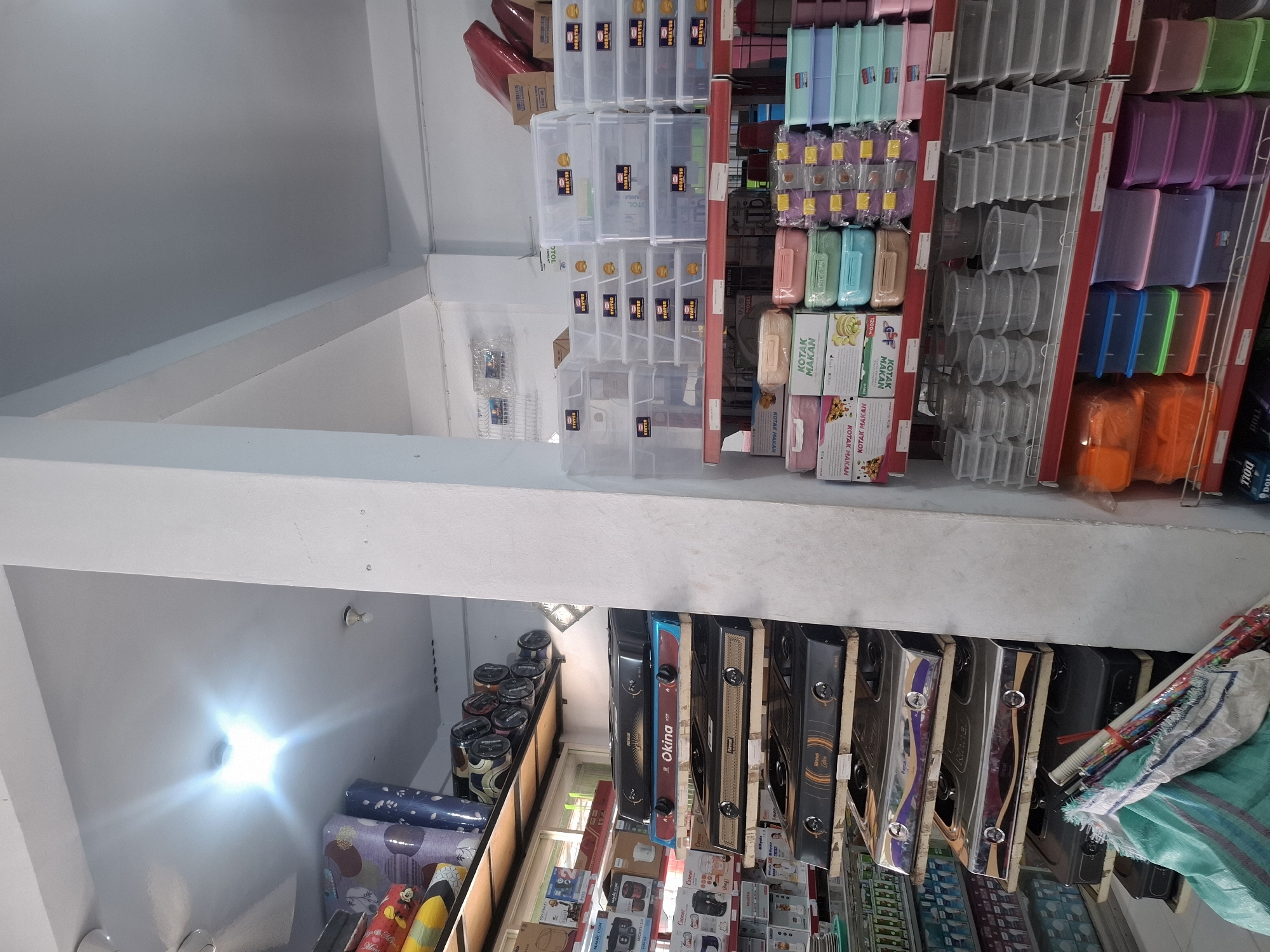 The image shows the interior of a retail store located in Sumberwudi, Indonesia, likely a home goods or kitchenware shop. The store is well-stocked with various items, arranged on shelves. In the foreground, there are stacks of clear plastic storage containers of different sizes on a red shelf. To the left of the plastic containers, on a lower shelf, are colorful food storage containers. Above these, on wooden shelves, are several gas stoves, stacked one on top of the other. The brands "Rinnai" and "Okina" are visible on the stoves. Further to the left, on higher shelves, are rolled items that appear to be carpets or mats, along with boxes and smaller appliances. The lighting in the store is bright, with some glare from overhead lights. A white pillar divides the space. A pile of bundled fabric or plastic sheeting is visible in the lower left corner of the image, obscuring some of the lower shelves.