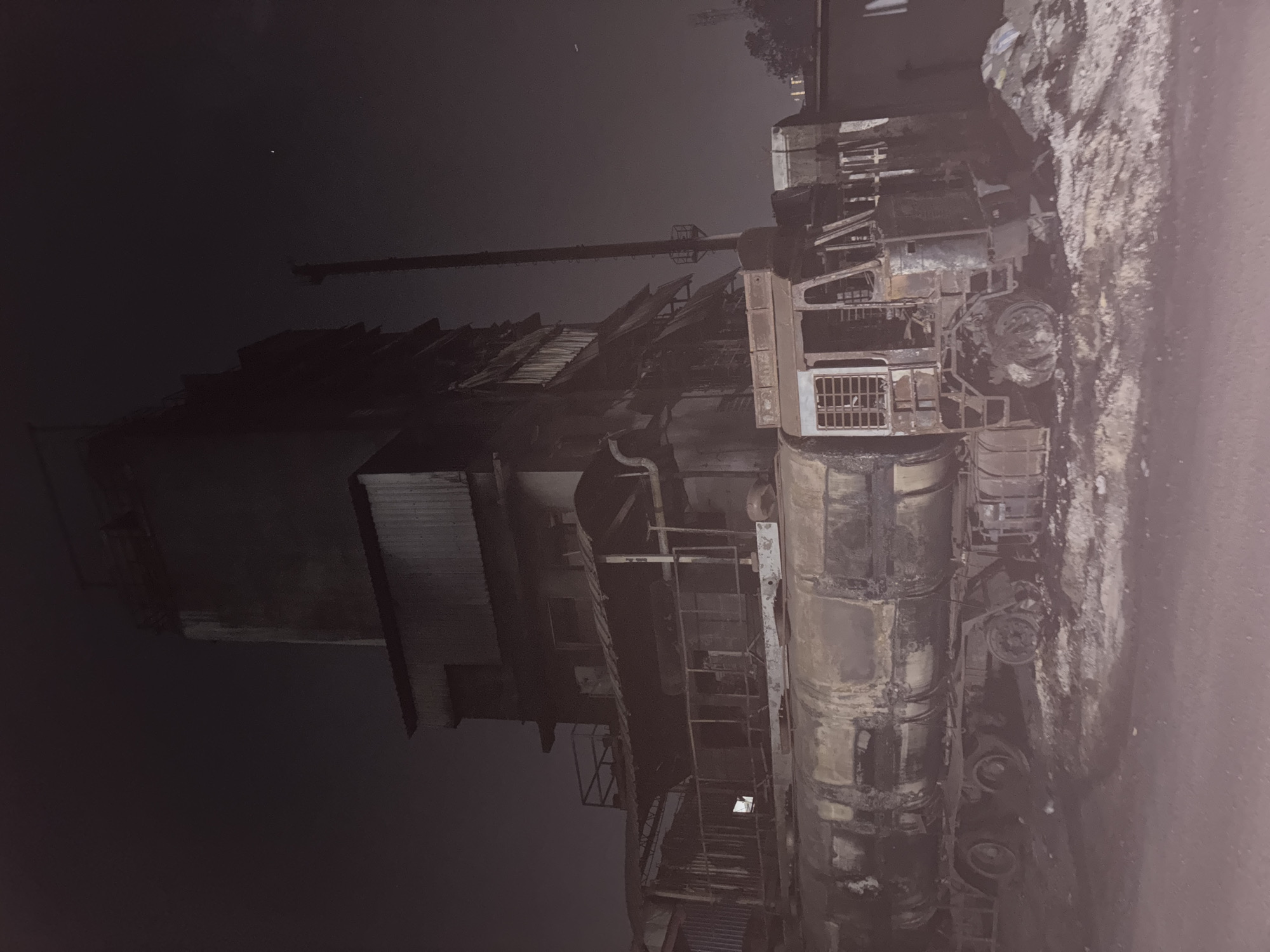The image shows a severely damaged industrial facility at night. Charred remains of what appears to be a truck are visible on the right side of the frame, indicating a possible explosion or fire. The building structures are heavily damaged and marked with soot. A tall tower stands on the left side, and other industrial equipment can be identified. The location is potentially Ambernath, India, based on the request.