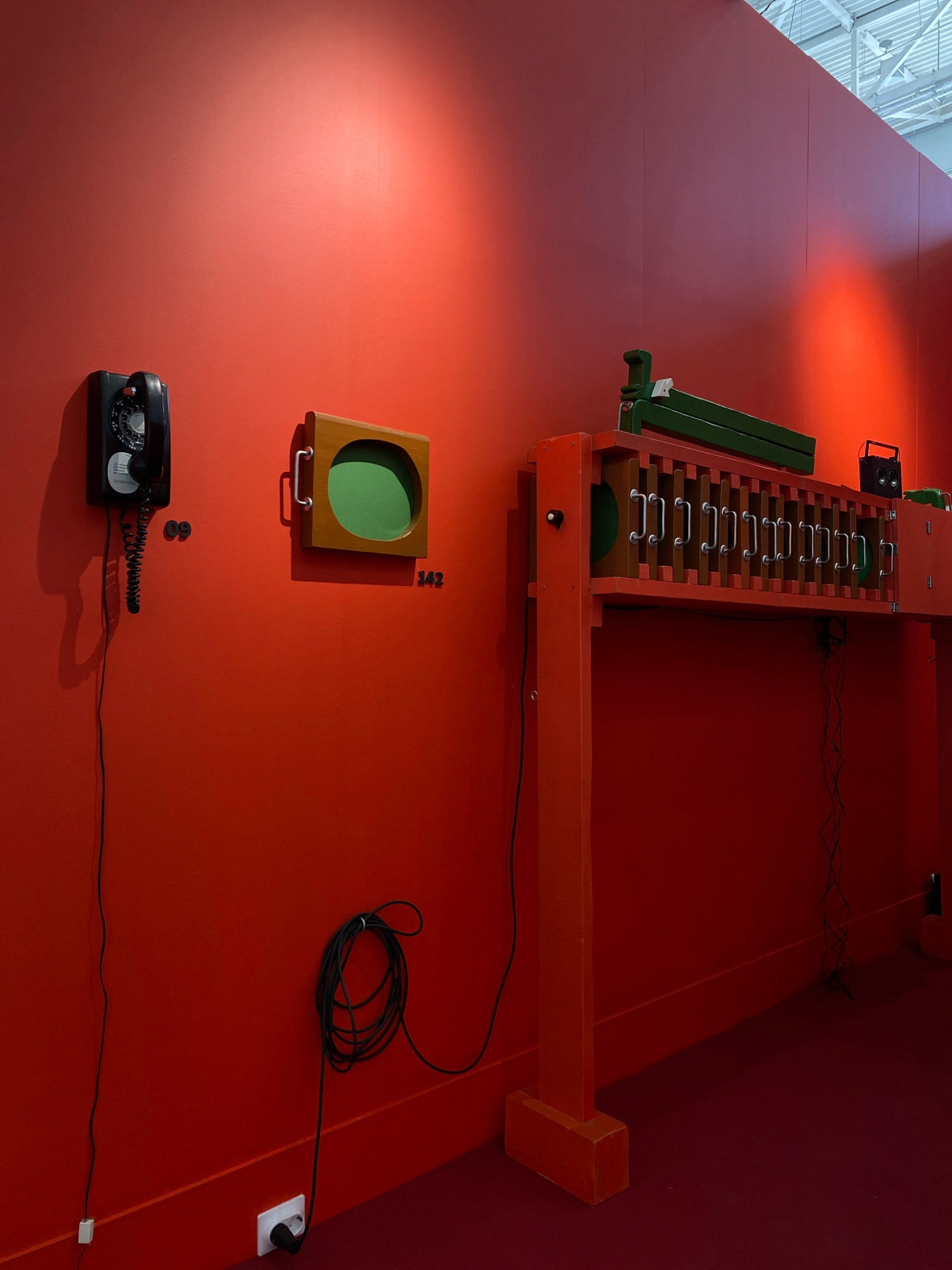 The image depicts an art installation on a vibrant red wall in a museum or gallery space, possibly located in Khimki, Russia. The installation features a vintage black rotary telephone mounted on the left side of the wall, with its coiled cord hanging down. To the right of the telephone, a square wooden frame with a green interior is mounted on the wall. Further to the right, a large, orange, multi-part wooden structure dominates the scene. This structure has a top section with multiple metal handles, reminiscent of a large-scale musical instrument or an interactive art piece. A green element is placed on top of this structure, and a black speaker is attached to the right side. Black electrical cords are visible, connected to an outlet on the floor. The floor is covered in a deep red carpet. The lighting suggests artificial illumination from above, casting shadows on the red wall. There are no people visible in the image.