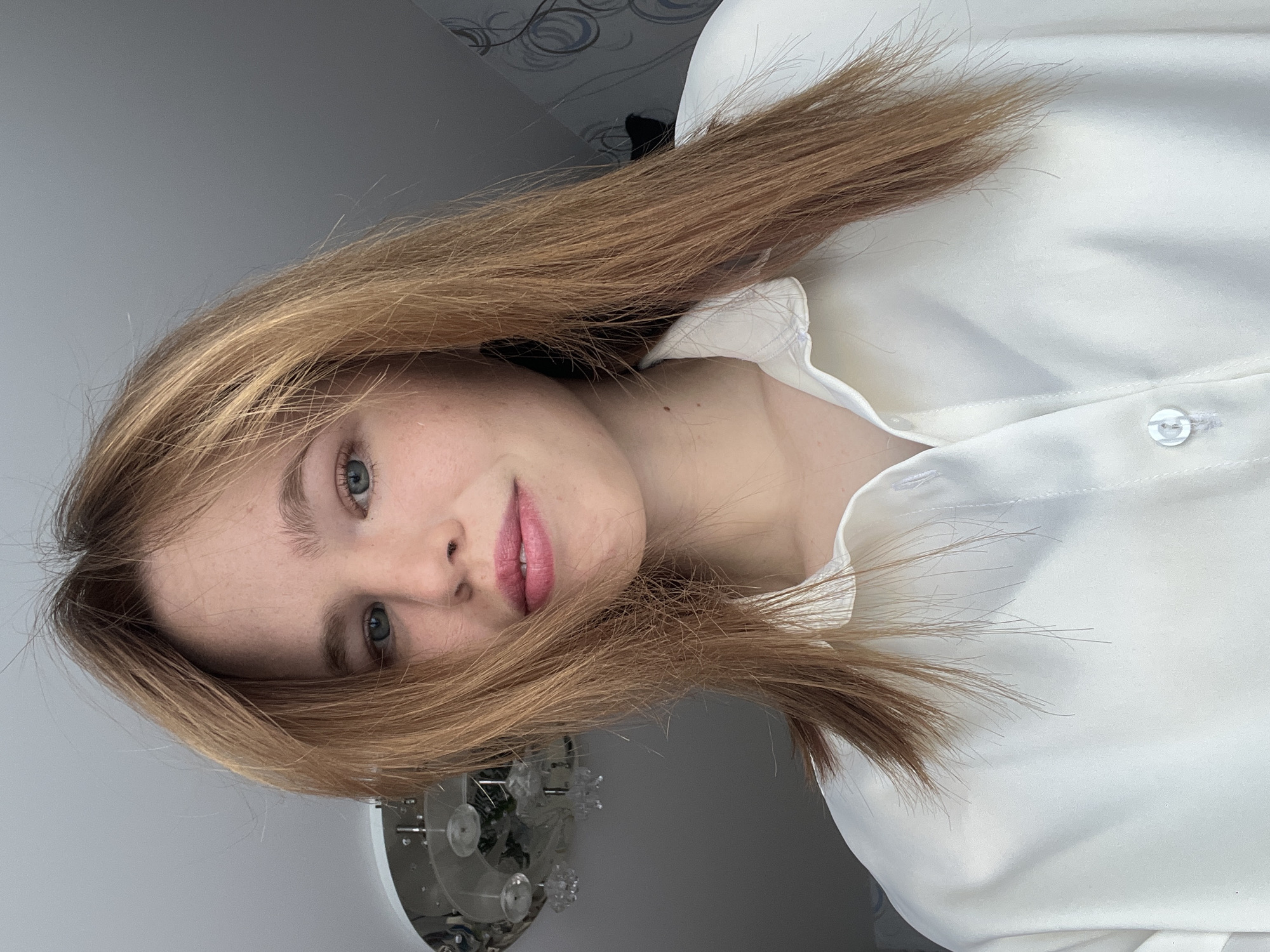 An indoor portrait taken in the City of Chaikovsky, Russia, features a young female subject looking directly into the camera. The subject has fair skin, light blue eyes, and reddish-blonde hair parted roughly in the middle, extending past her shoulders. Visible freckles are present on her nose and cheeks. Her lips are adorned with pink gloss or lipstick. She is wearing a plain, collared, long-sleeved white or off-white button-down shirt, with the top button fastened. The background consists of a light gray ceiling in the upper left, where a multi-bulb, decorative ceiling light fixture with clear or frosted elements is partially visible. To the right, a wall with a subtle, abstract gray and blue patterned wallpaper is discernable. The lighting is consistent with an indoor setting.