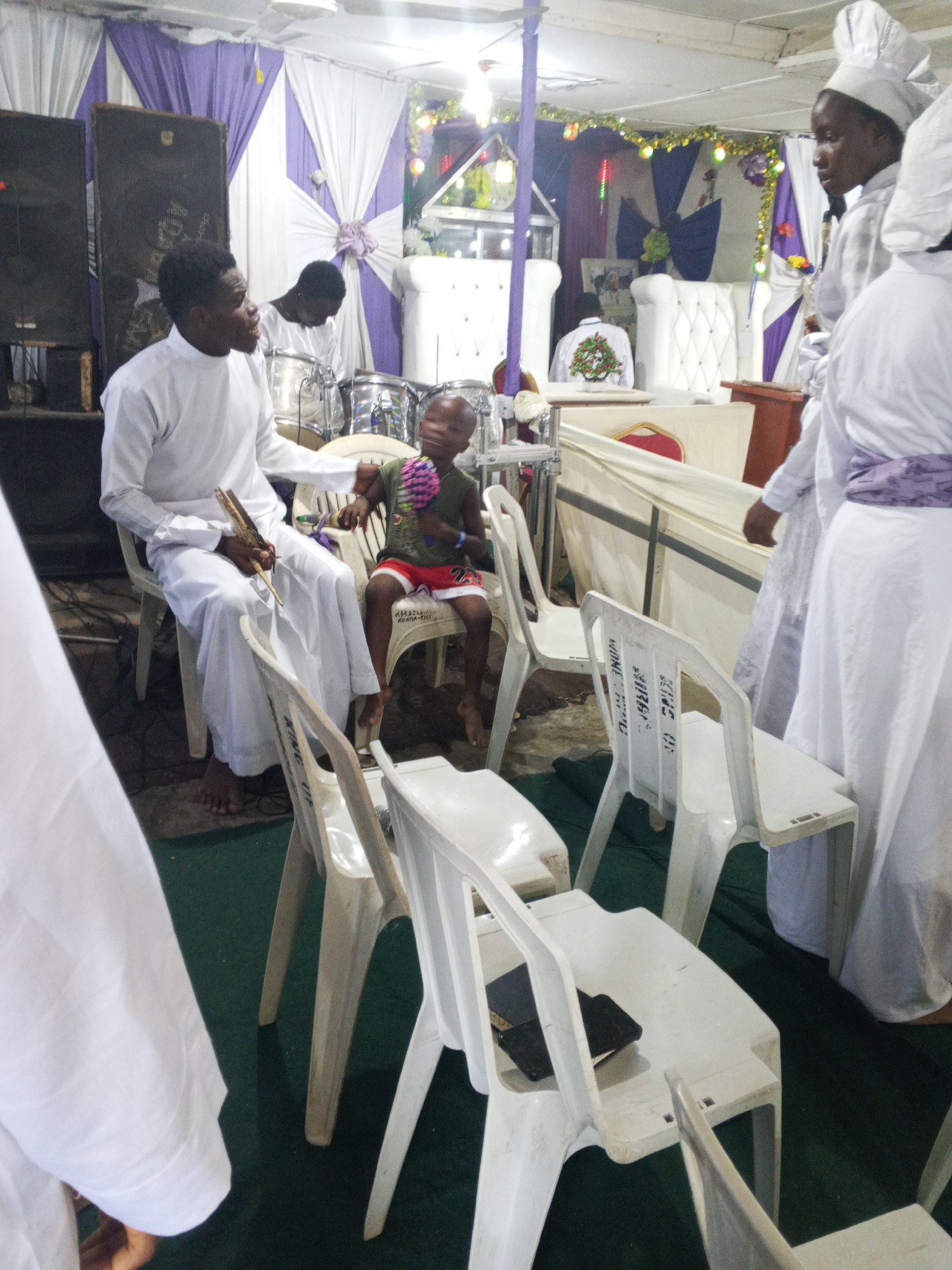 The image depicts a scene likely within a church setting, recognizable by the presence of individuals in white garments. A man in a long white robe, holding what appears to be musical sticks, sits next to a child on a chair who is holding colorful noise instruments. Another man in white sits behind drums, and there are several other people around. The scene is set indoors under a white curtain, with a decorative arrangement in the background. White plastic chairs are visible in the foreground. The location in Lokoja, Nigeria, is based on the information in a similar way as what would be written on the chair, a name of the church.