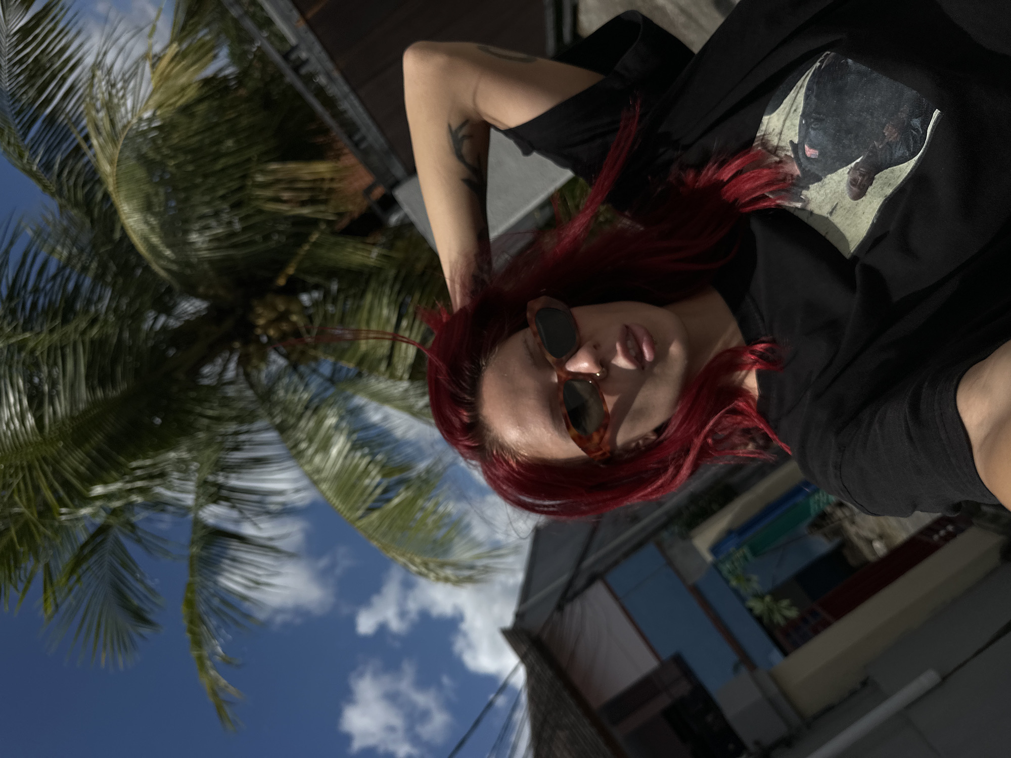 A woman with red hair and sunglasses is shown in a portrait-style photograph, with a palm tree and a blue sky with white clouds in the background. She is wearing a black t-shirt with a graphic on it. The location is likely in North Kuta, Indonesia, based on the tropical environment. Buildings are visible in the background. The woman has a tattoo on her left arm. She appears to be posing for the photo.