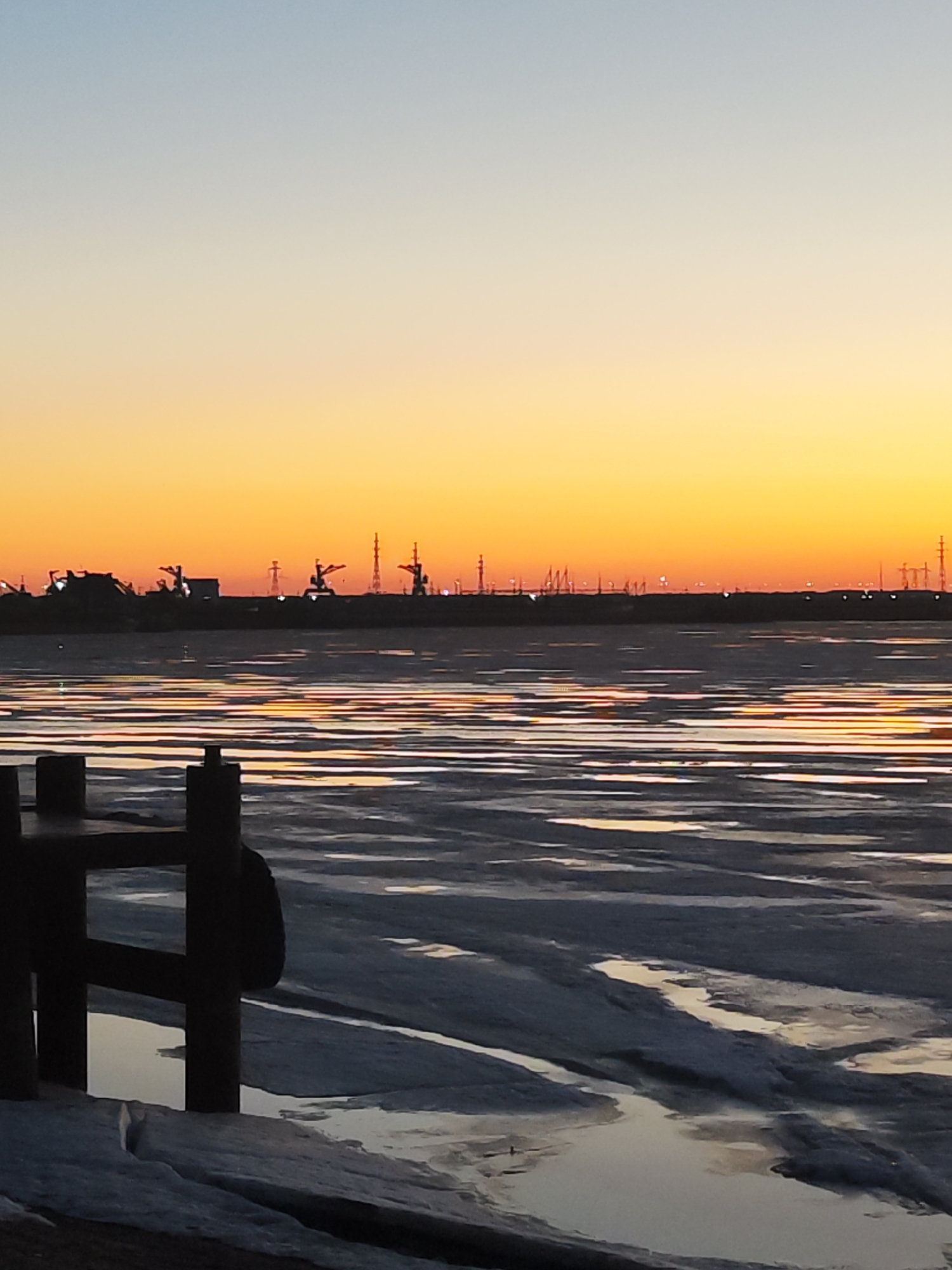The image captures a winter scene at sunset on the Kama River in Chaikovsky, Russia. The sky displays a gradient of colors, transitioning from a pale blue at the top to a vibrant orange near the horizon. The setting sun reflects on the partially frozen river surface, creating horizontal streaks of light. In the middle ground, a dark, silhouetted cityscape is visible, marked by industrial structures and power lines. In the foreground, a portion of a wooden pier or platform is present, its dark vertical posts and horizontal beams framing the view. The ice-covered ground near the pier shows patches of open water and snow.